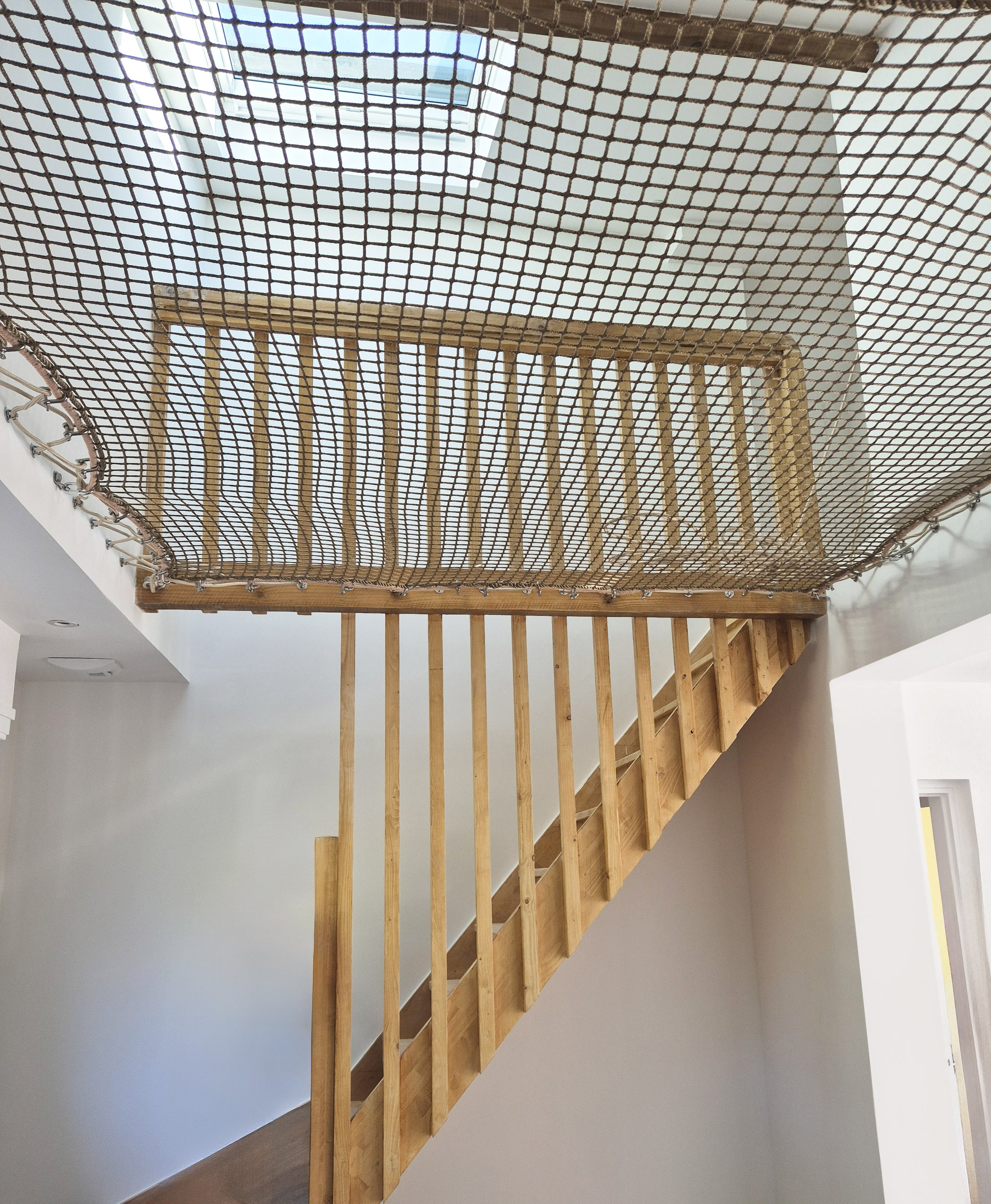 Escalier en bois avec rampe, vue depuis le bas, avec un filet de sécurité au plafond pour assurer la sécurité.