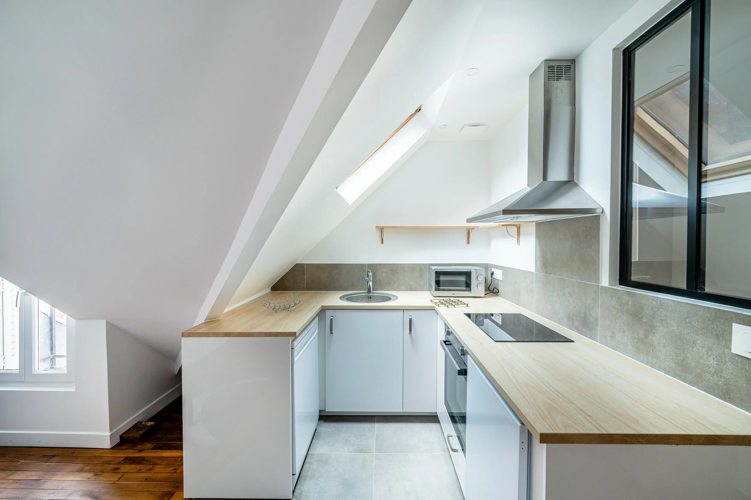 Cuisine moderne avec comptoir en bois, évier, micro-ondes, cuisinière électrique, hotte en acier inoxydable, fenêtre et plafond en pente avec un velux.