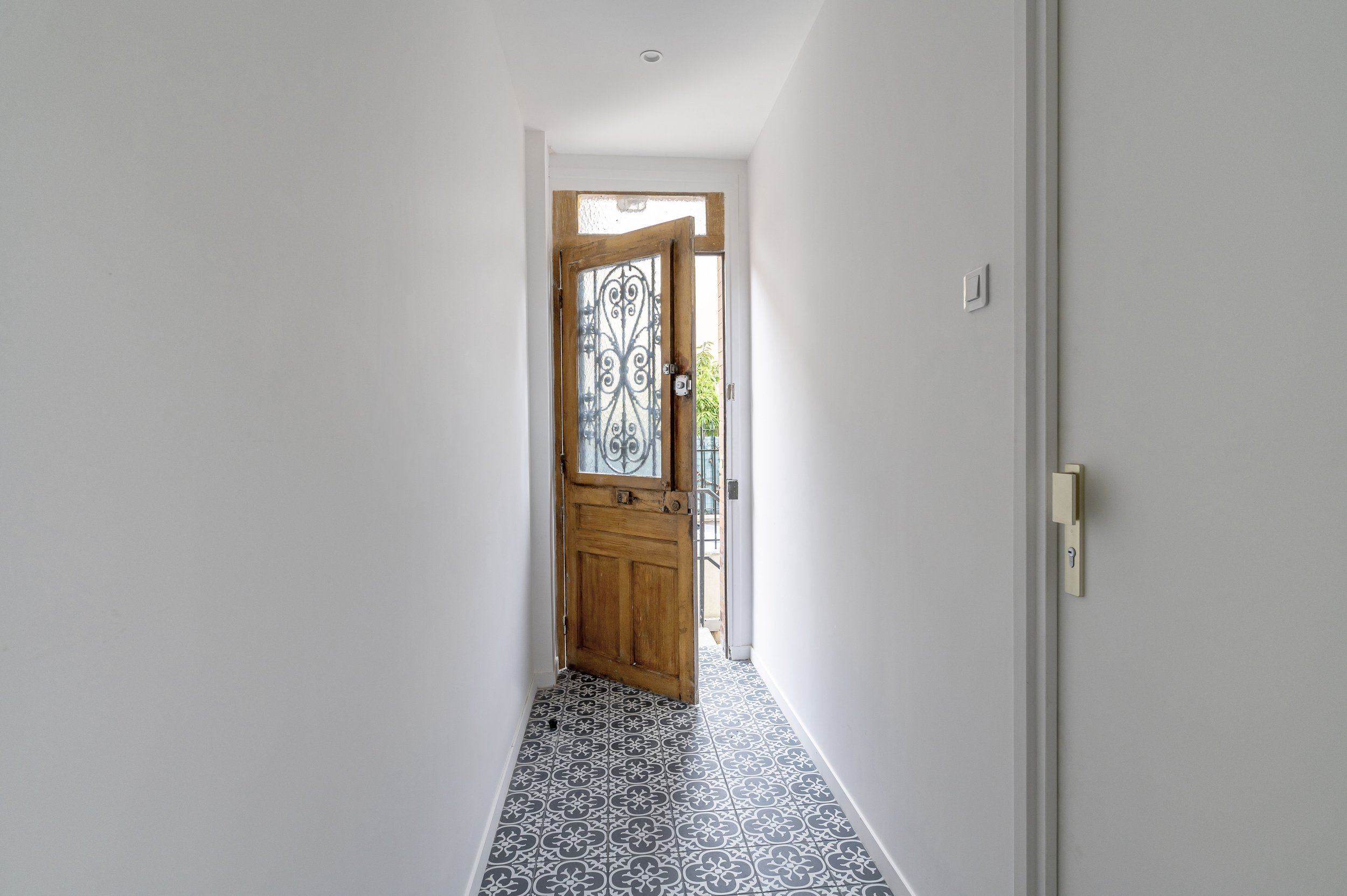 Vue d'un couloir avec porte d'entrée en bois et vitrail, sol en carreaux noirs et blancs, murs blancs, lumière naturelle.