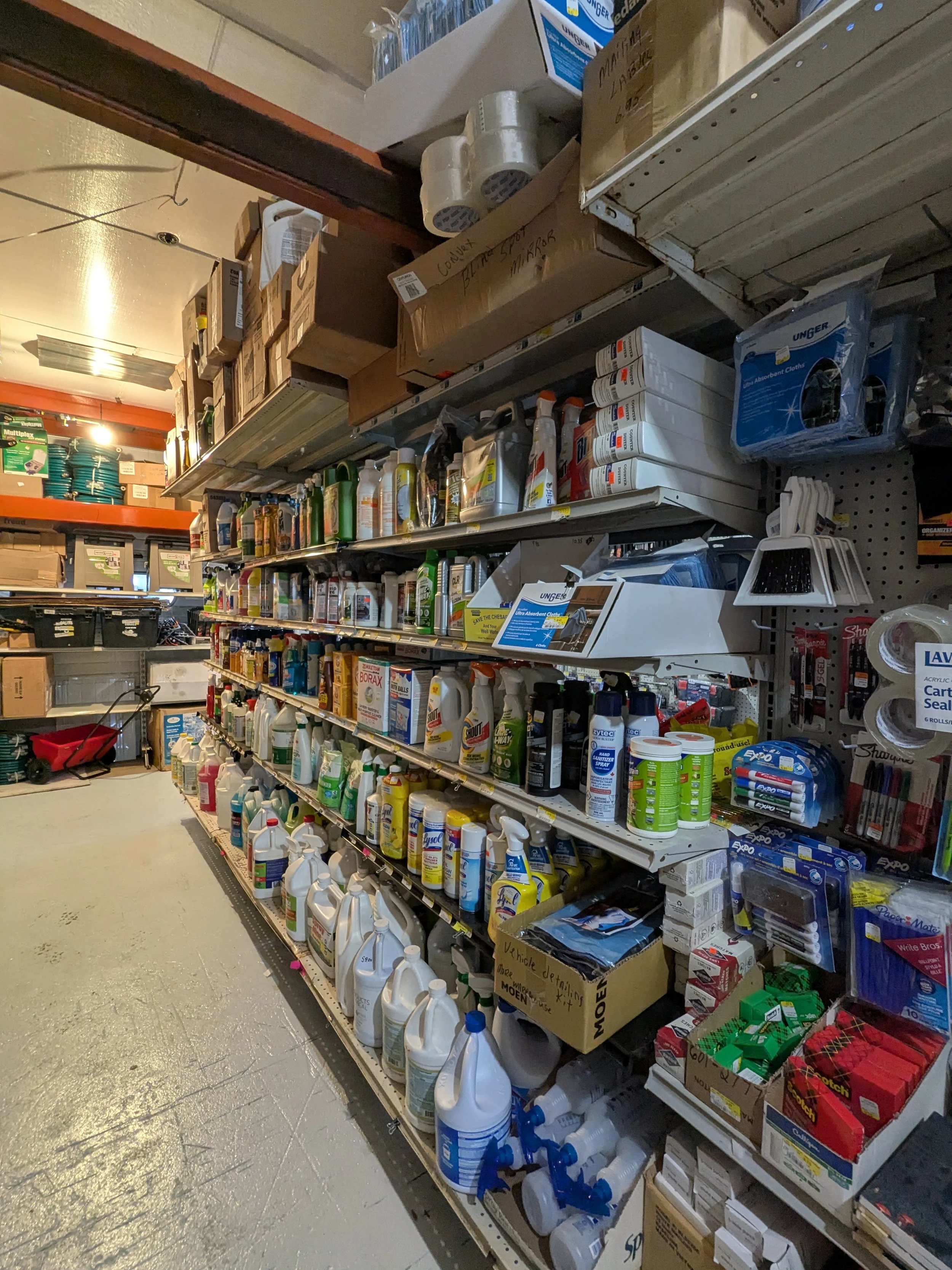 Aisle in a hardware store with shelves stocked with cleaning supplies, sprays, bottles, and boxes of assorted products.