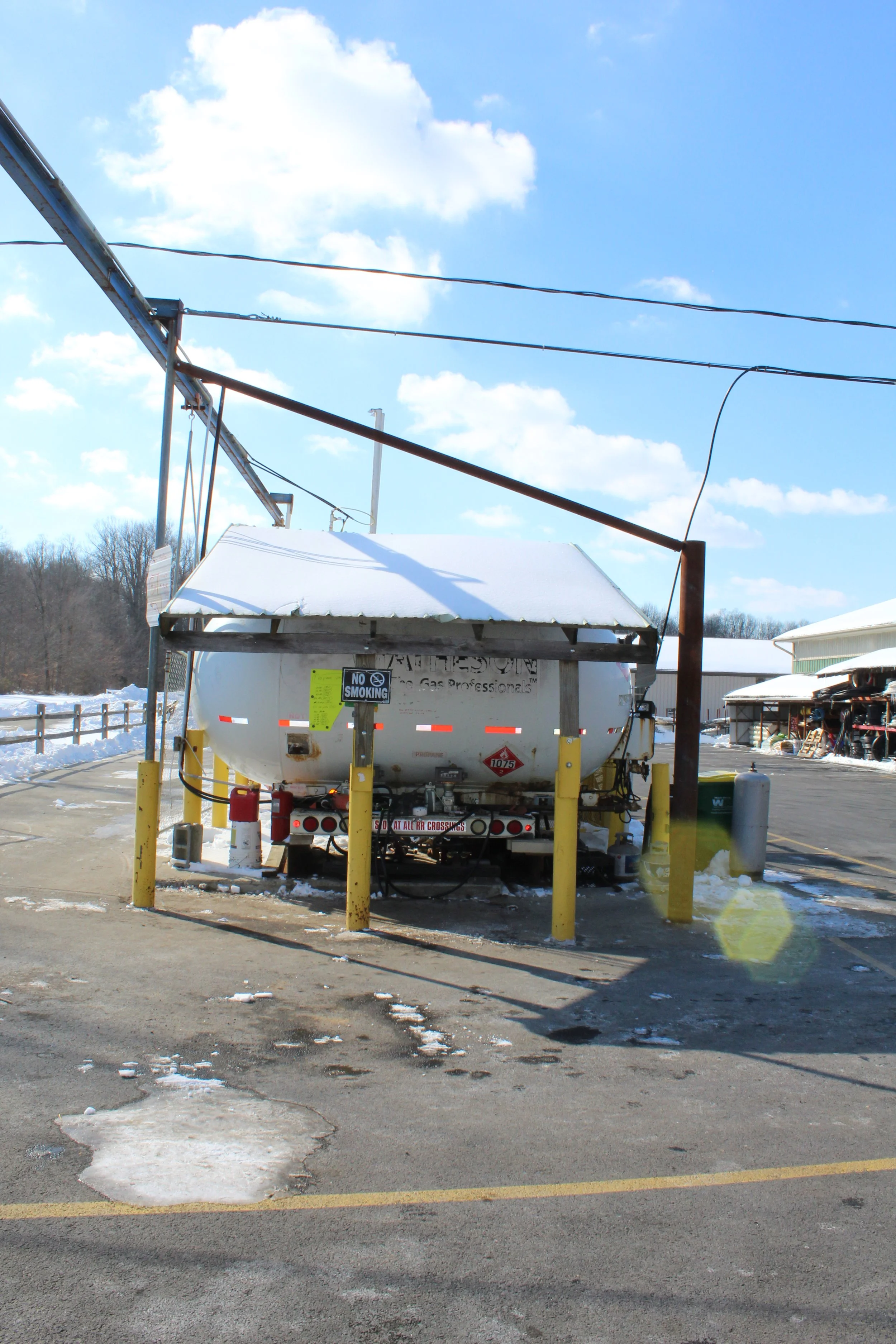 A propane tanker truck parked in a snowy area with a canopy, surrounded by yellow posts, under a blue sky with clouds.