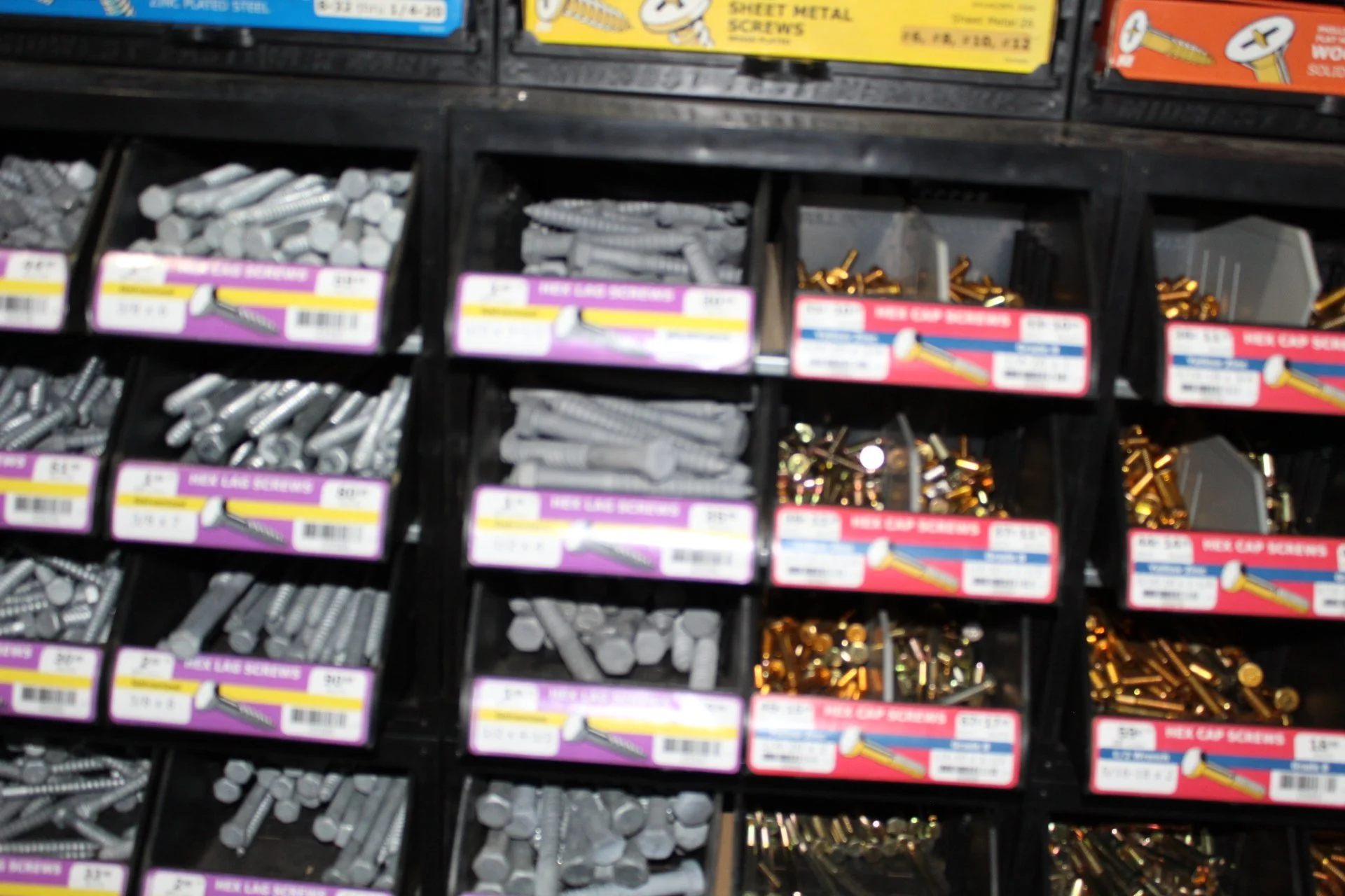 Blurred image of a retail hardware store display with compartments holding various screws and fasteners, including silver, gold, and brass-colored screws.