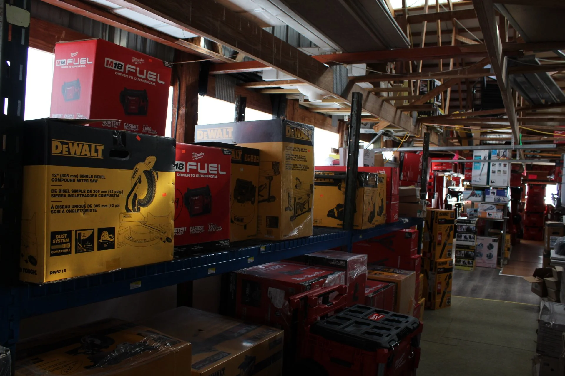 A hardware store aisle filled with power tools and equipment in boxes, including a DeWalt mitre saw and Milwaukee fuel tools, with shelves and stored items overhead.