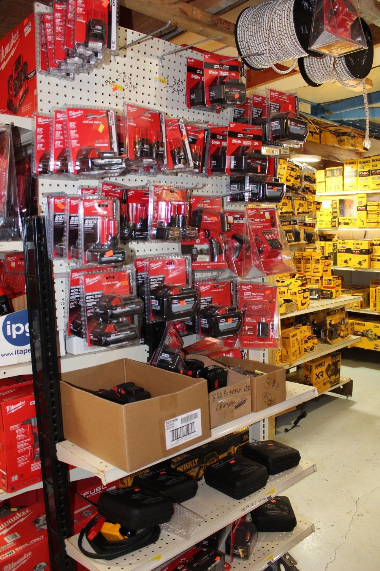 Power tools and accessories on store shelves, including Milwaukee cordless drills, batteries, chargers, and saws, with yellow DeWalt power tools in the background.