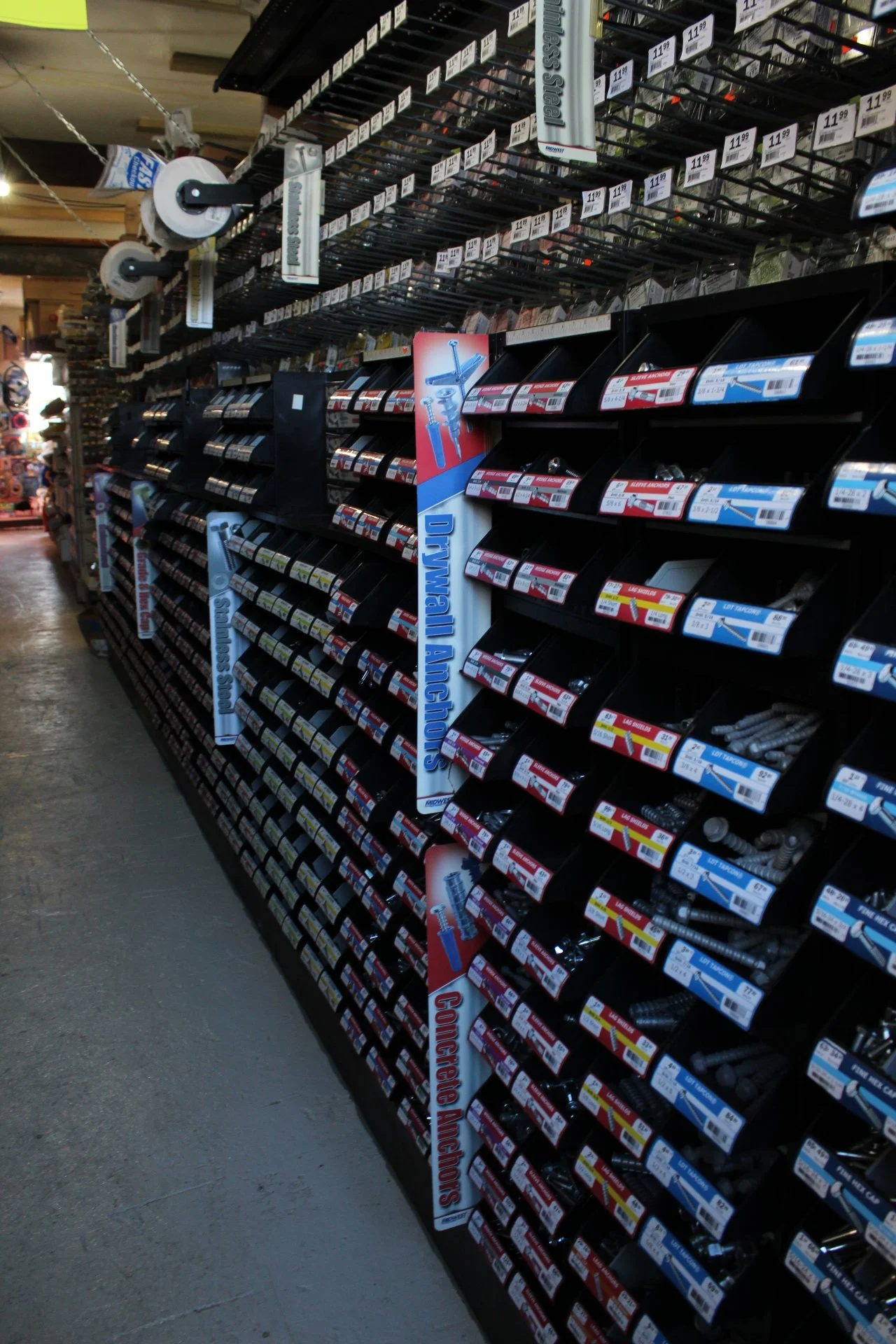 A hardware store aisle displaying screws and nails organized in black plastic trays with labels, with signs indicating sections for 'Bit Hardware' and 'Concrete Anchors'.