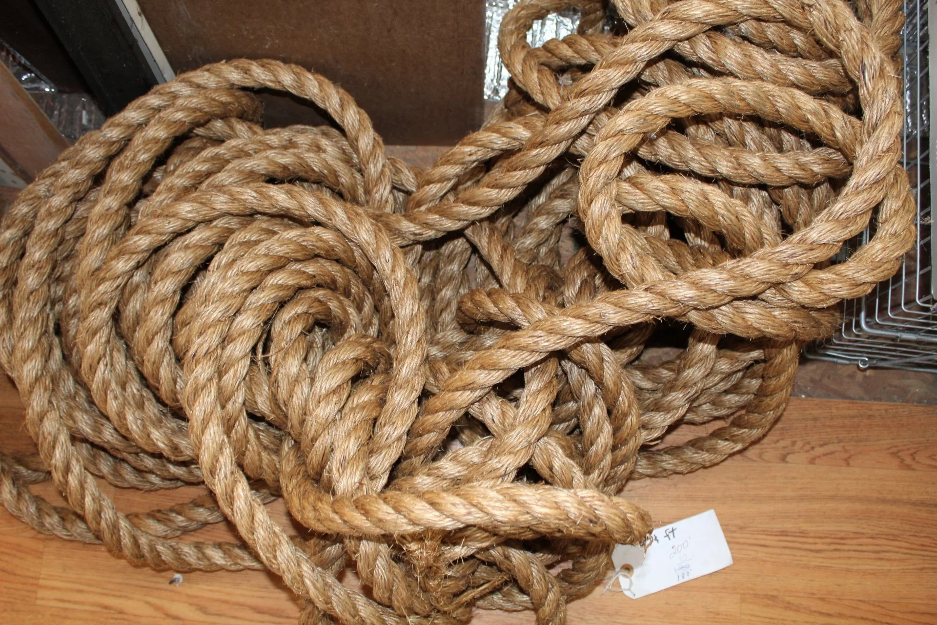 A pile of coiled thick, twisted rope made of natural fiber on a wooden floor, with a small piece of paper showing some handwritten text and a part of a metal object in the background.