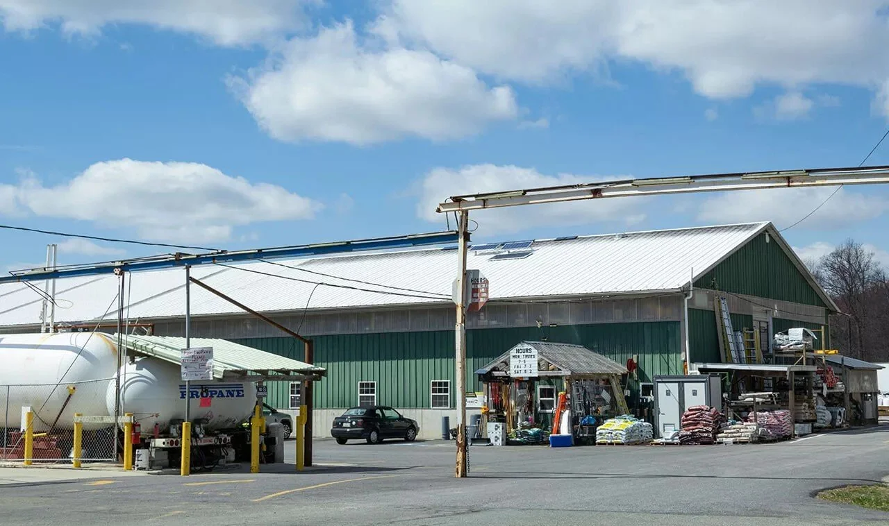 A large metal warehouse building with a green exterior, white metal roof, and small window. Outside the building are various items, including bags of fertilizer or soil, a small display shed, and equipment. A propane tank is parked nearby, and there are parked cars and a parking lot in the foreground. The sky is partly cloudy.