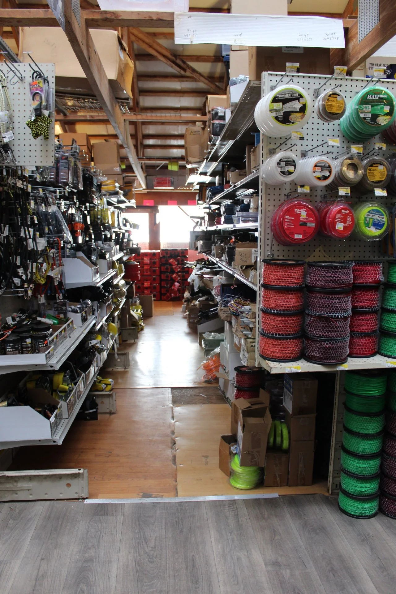 Shelf in a hardware store aisle with various tools and supplies, including extension cords, tape measures, and storage containers, with a bright exit at the back.
