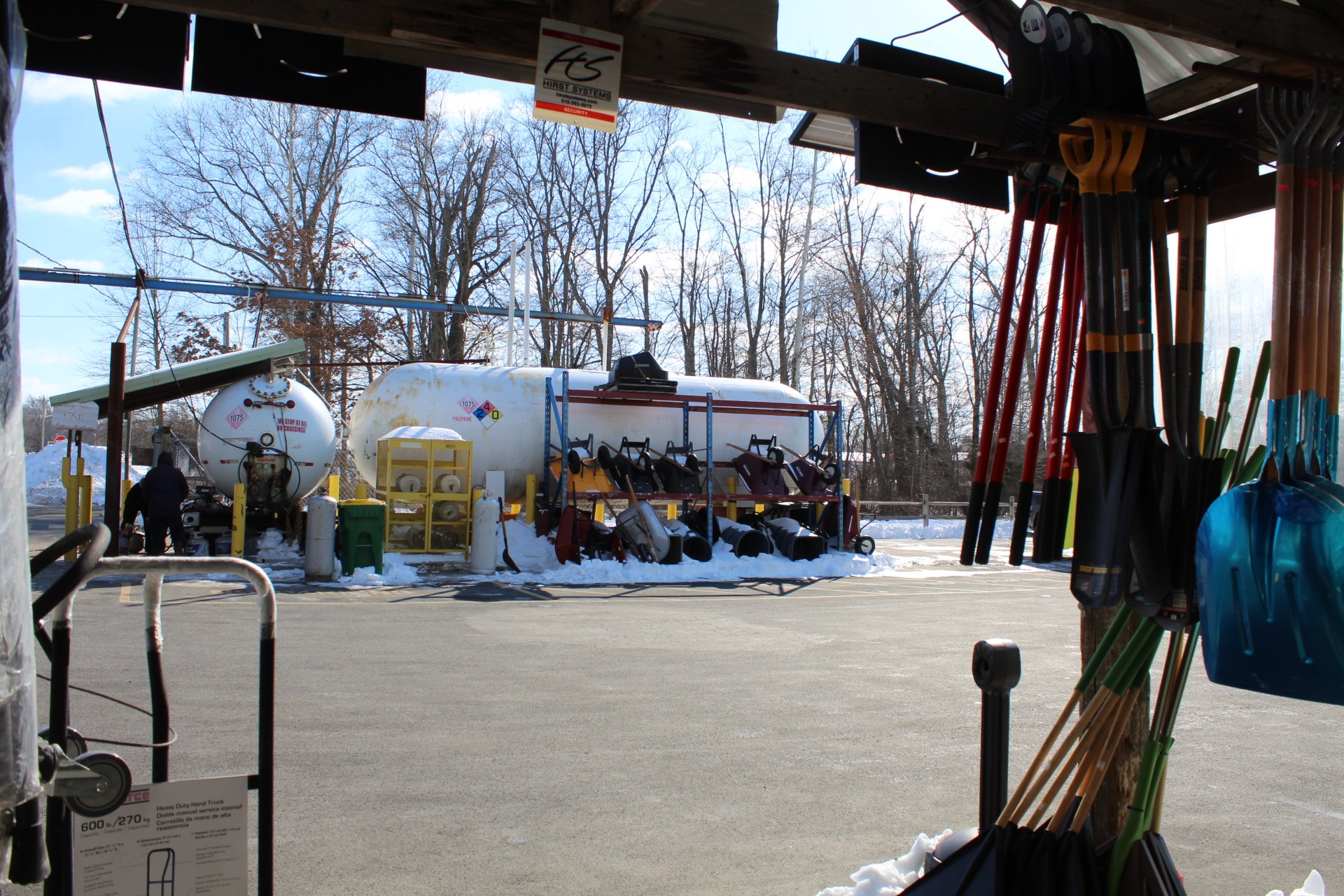 View from a hardware store looking out onto the parking lot with snow on the ground, showing snow shovels, salt spreaders, and propane tanks, with leafless trees in the background and clear blue sky.