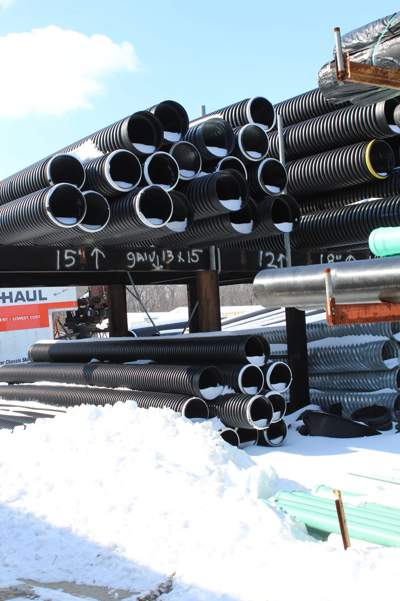 Stacks of large black plastic pipes stored outdoors on a metal rack, with snow on the ground and a blue sky in the background.