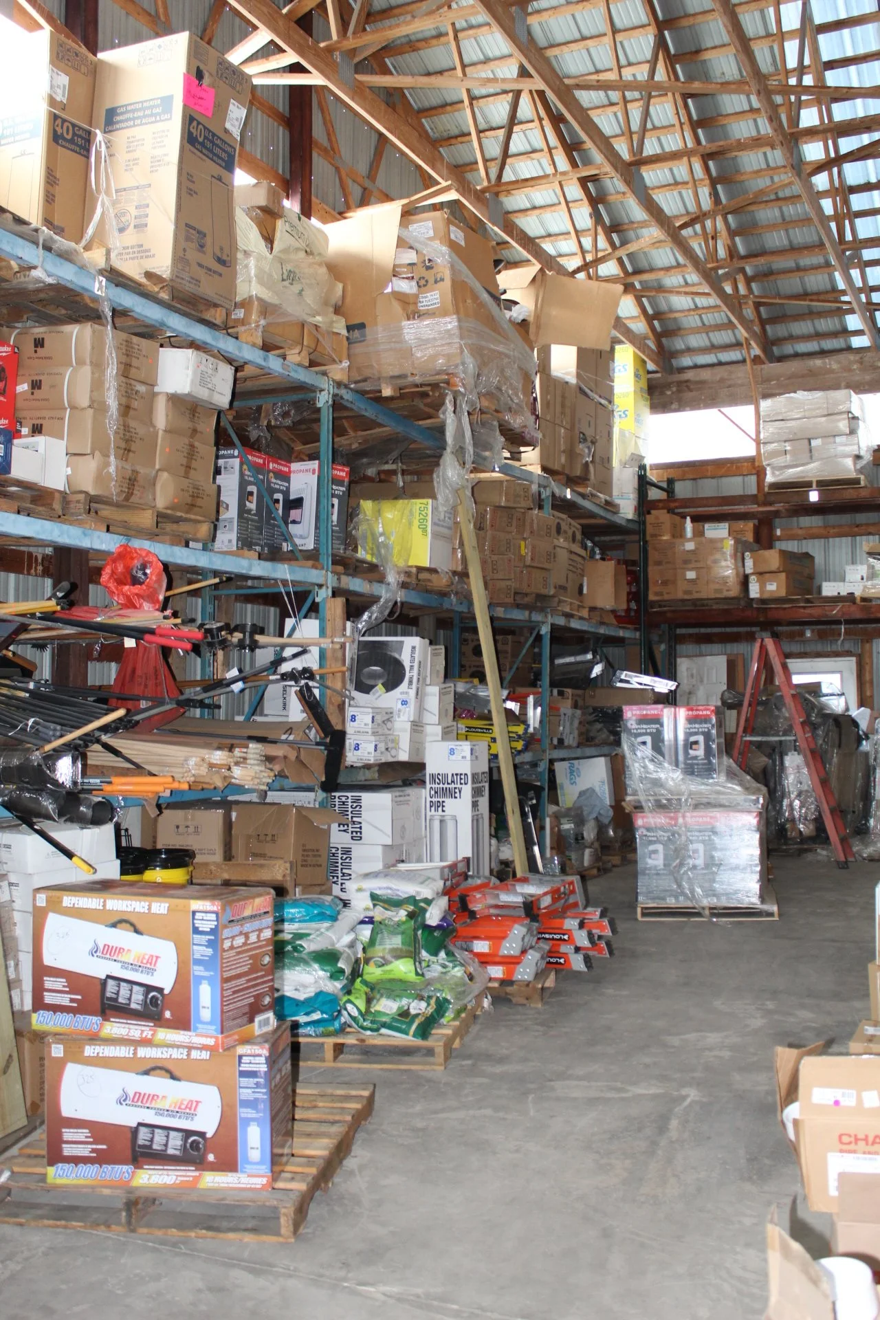 Shelves stocked with boxes of various sizes, some opened, in a warehouse with a metal roof and concrete floor.