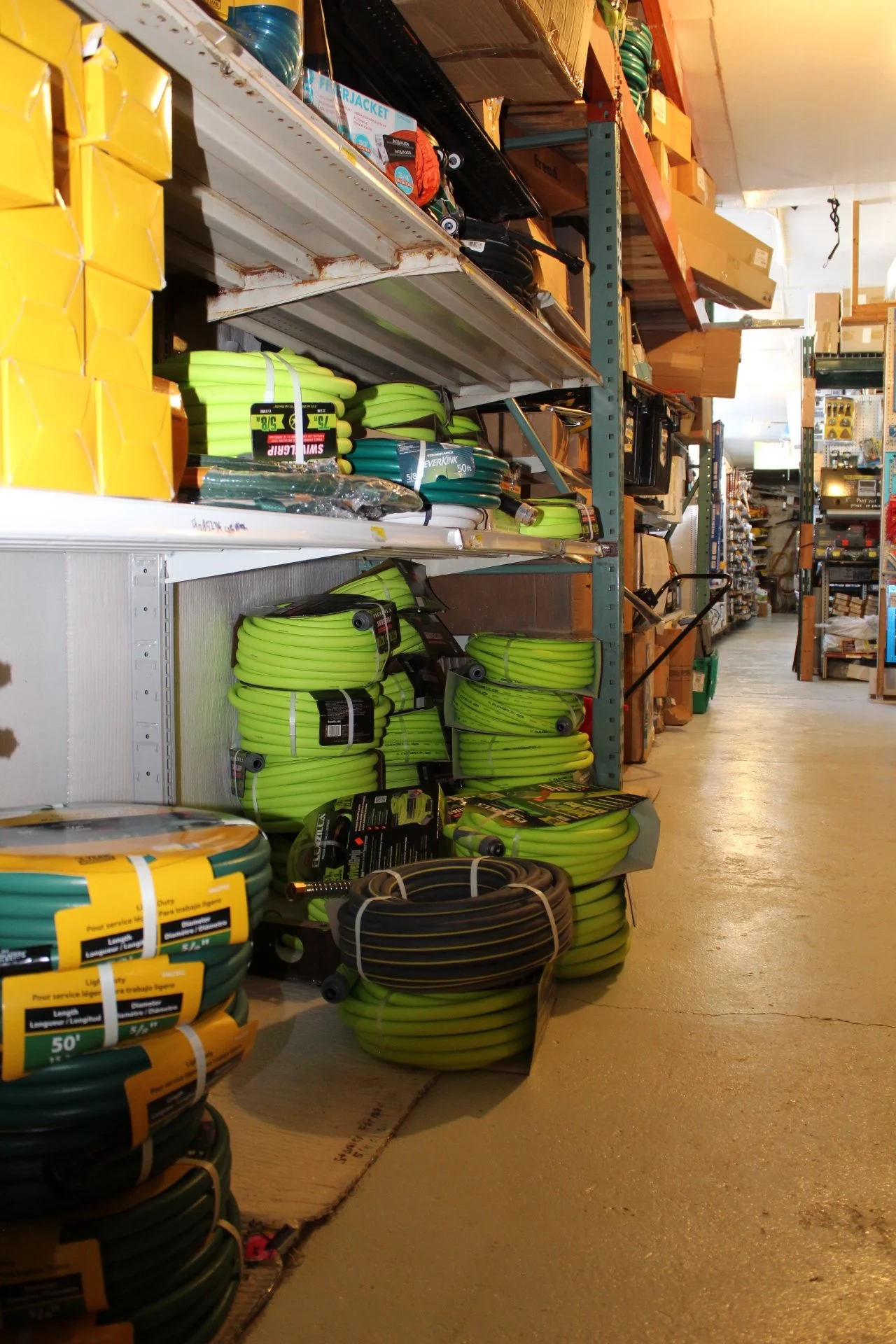 Store aisle with shelves stocked with various gardening supplies, including coiled green and black hoses, boxes, and garden tools.
