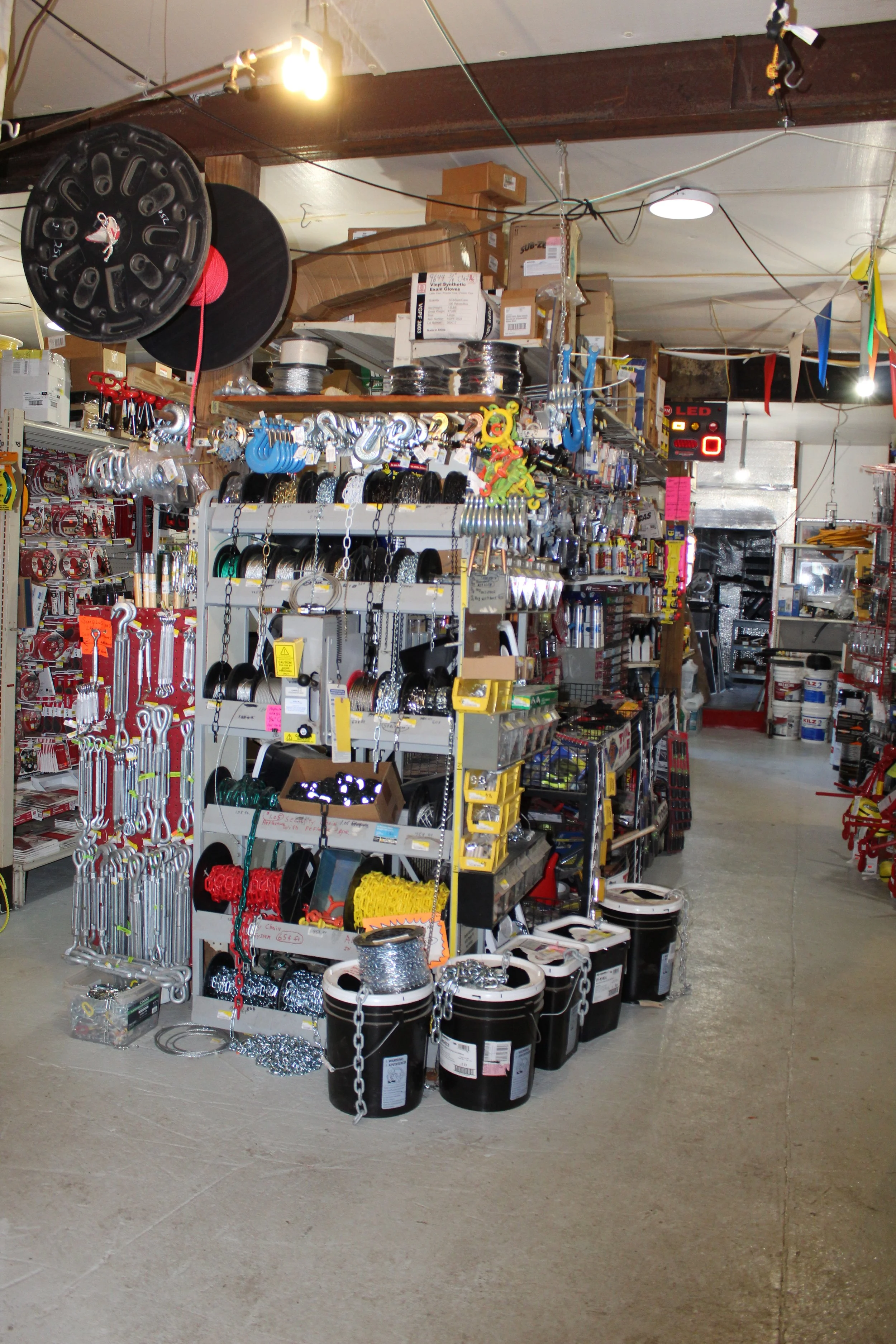 Hardware store aisle with tools, chains, rolls of wire, and buckets on display.
