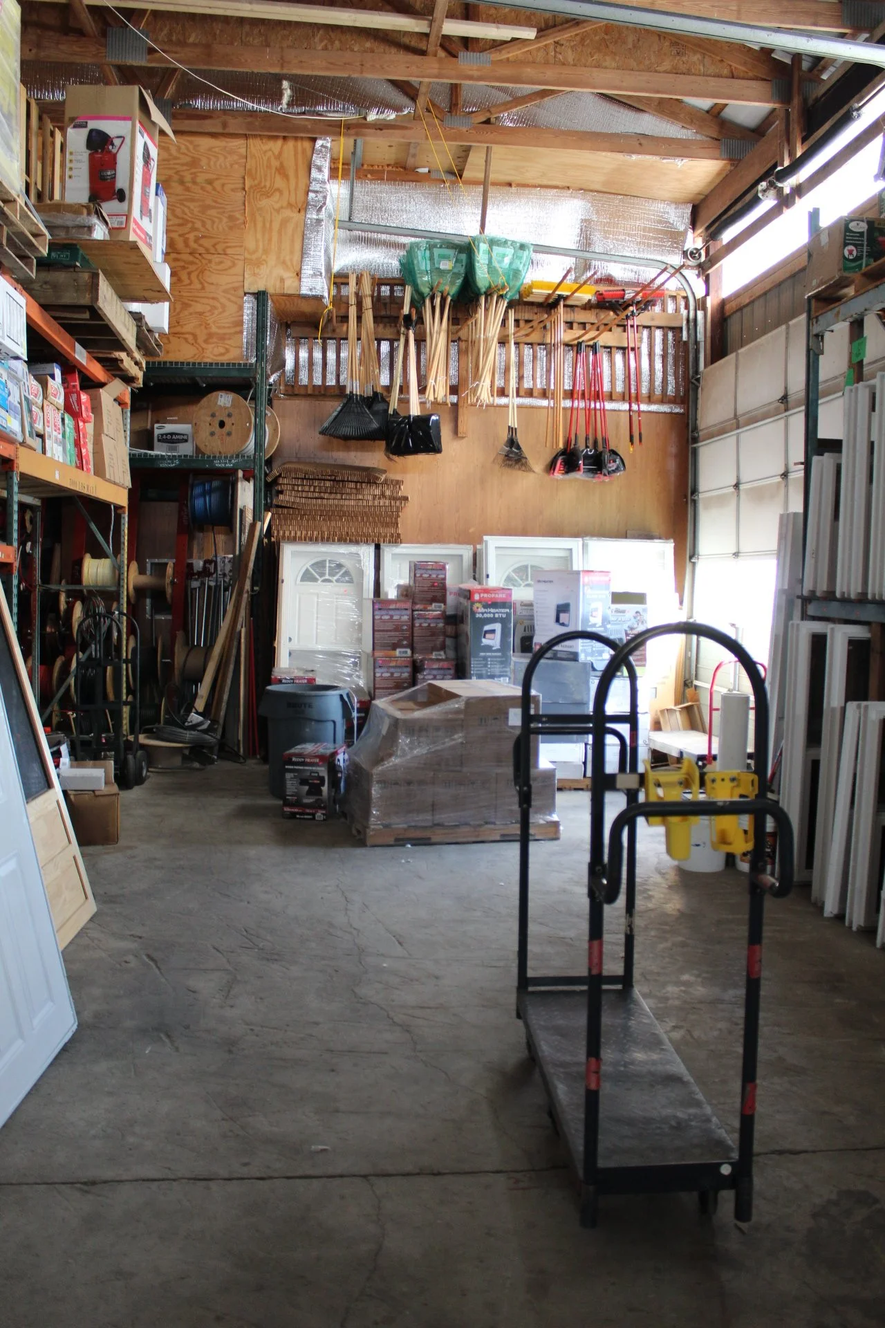 A cluttered garage with various tools and equipment, including brooms, rakes, and shelving units filled with boxes and supplies.