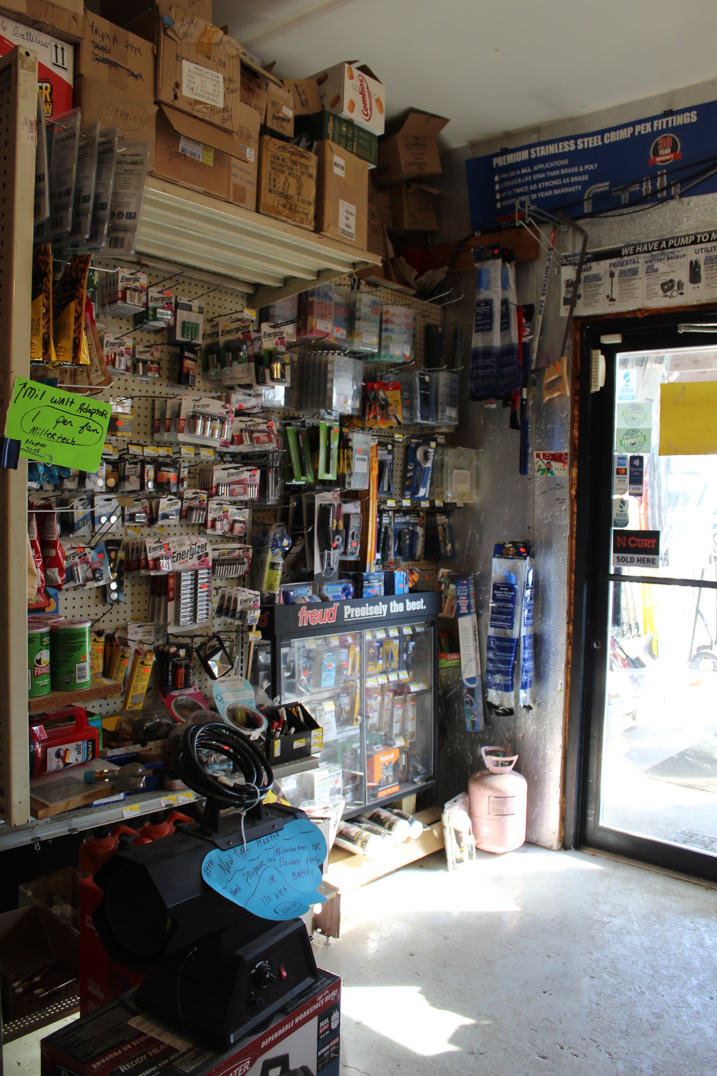 Inside a hardware store with shelves stocked with batteries, tools, and various hardware supplies near the entrance, with sunlight coming through the door.
