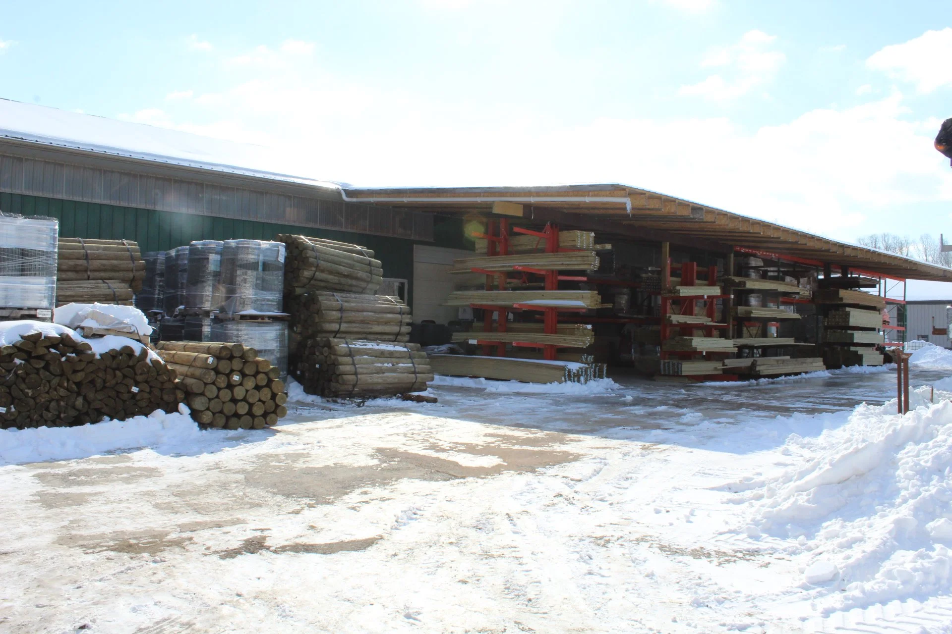 Outside view of a warehouse or lumber yard with stacks of wood and snow on the ground, under a partly cloudy sky.