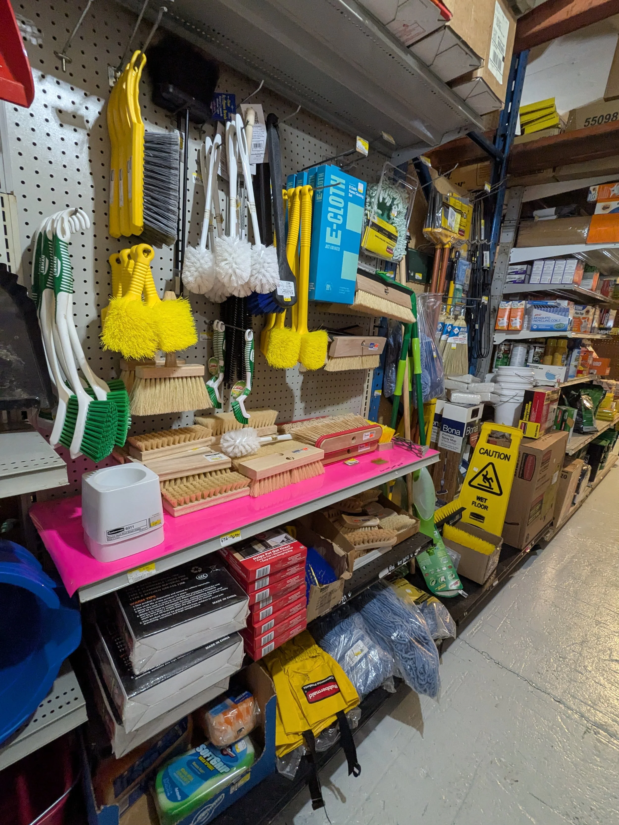 Store aisle shelf with cleaning supplies including scrub brushes, push bads, and mops, along with cleaning chemicals and floor caution sign.