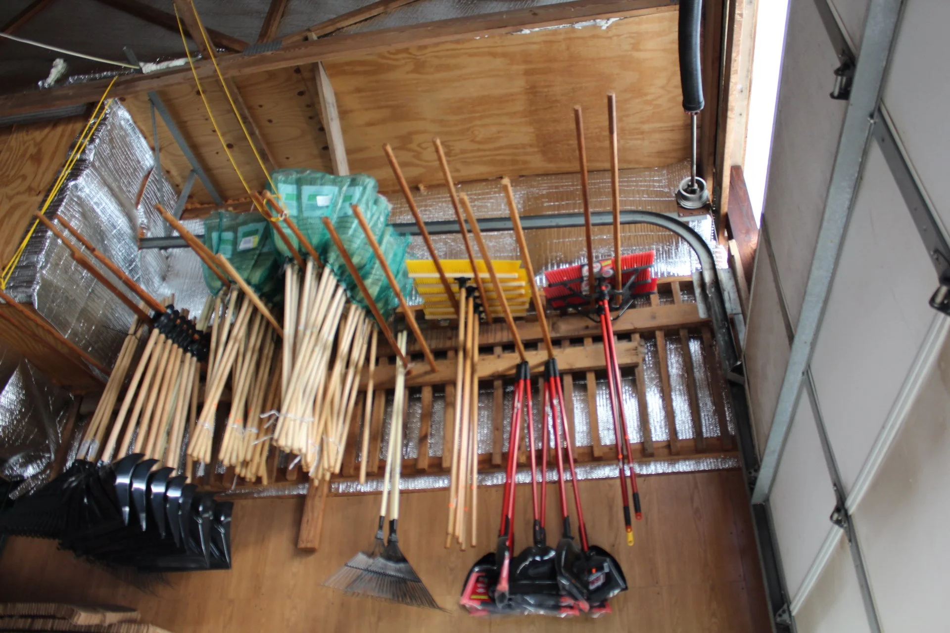 Garage storage area with brooms, shovels, rakes, and other gardening tools organized on a shelf and hanging on a wooden rack, with a garage door on the right.