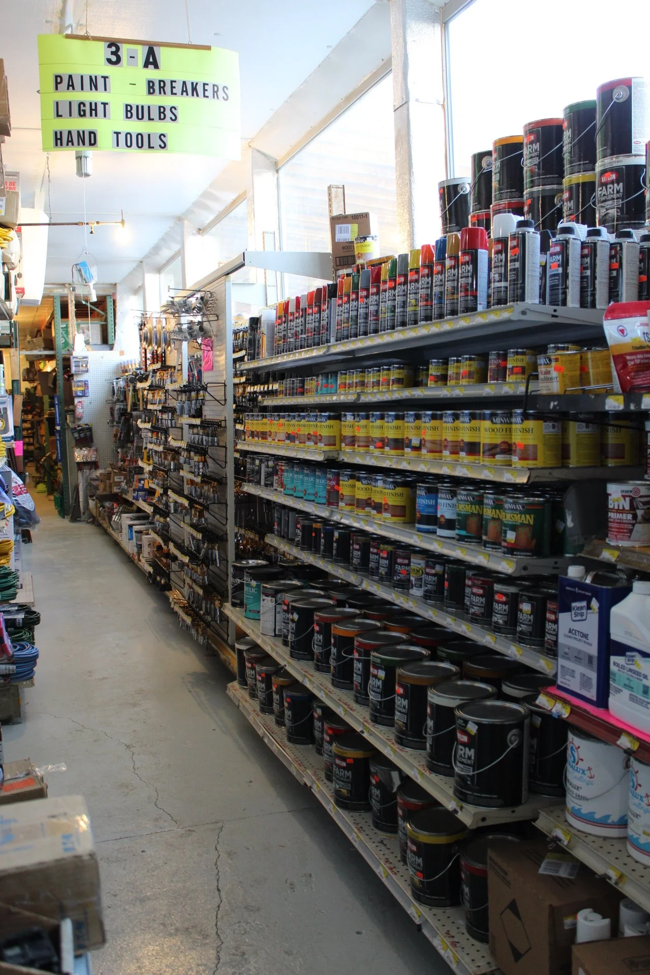 Aisle of a hardware store with paint cans on the right shelves and paint brushes on the left shelves, with a sign overhead labeled '3-A' indicating paint, breakers, light bulbs, and hand tools.