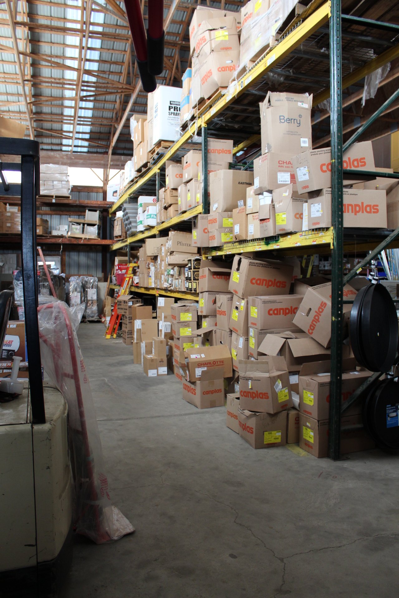Warehouse aisle with shelves stocked with cardboard boxes and equipment, with a concrete floor, metal roof, and natural light