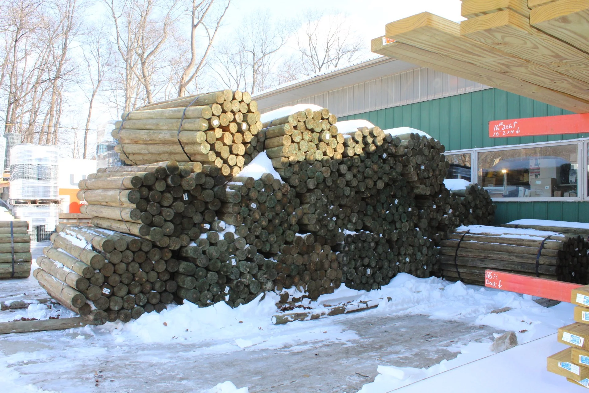 Stacks of wooden logs stored outdoors on a snowy day near a green building with a red marker indicating measurements.