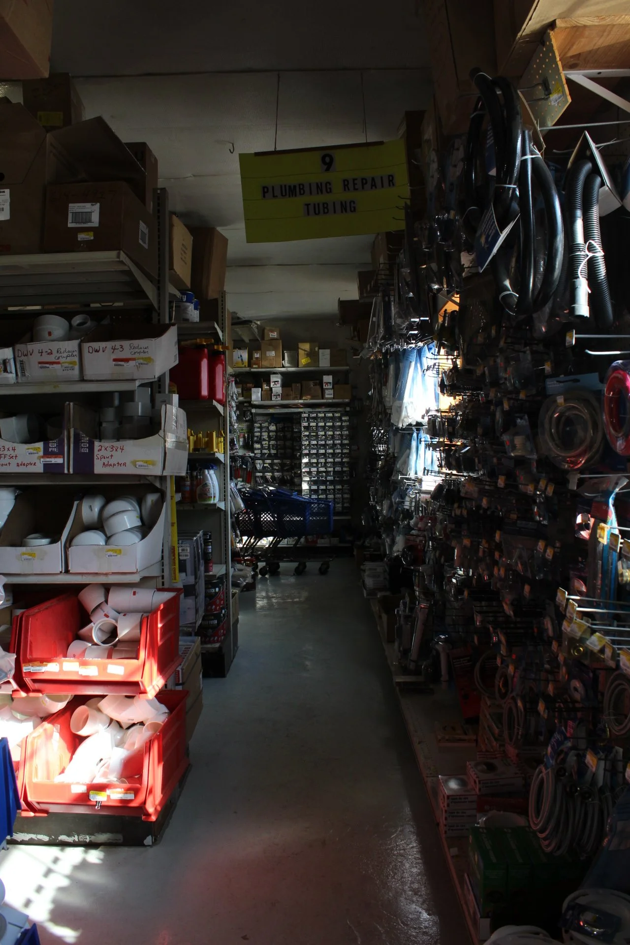 A hardware store aisle with plumbing supplies such as pipes, adapters, and hoses on shelves. A yellow sign reads 'Plumbing Repair Tubing' hanging above.