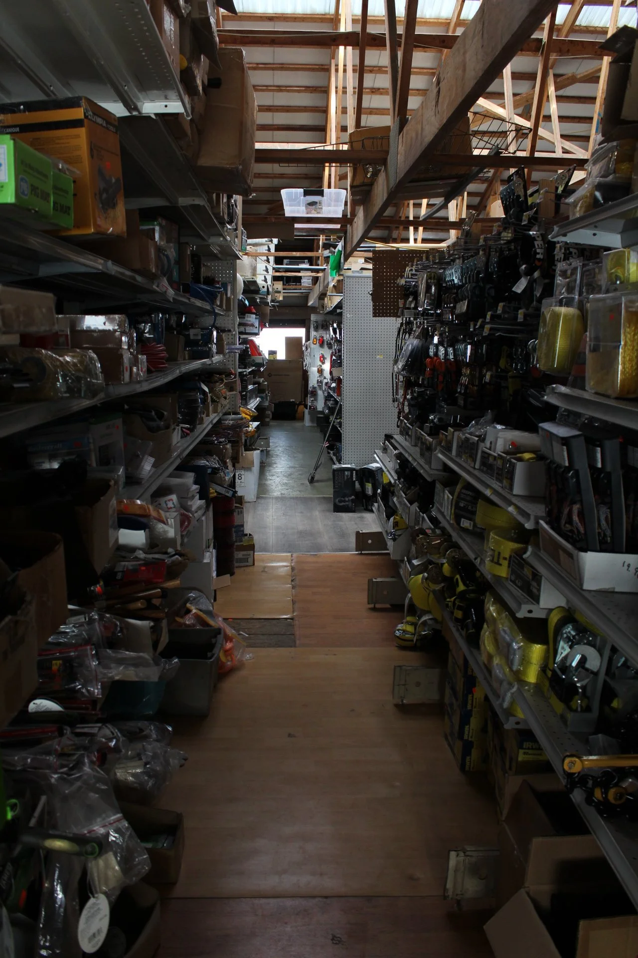 A warehouse aisle with shelves filled with various tools, hardware, and supplies, and a wood and metal ladder in the background, with sunlight coming through the windows.