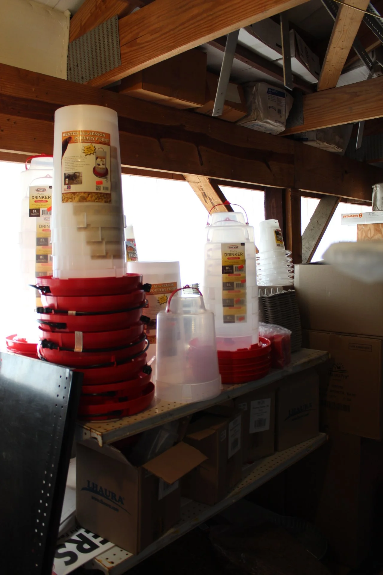 Stacked red and clear plastic poultry waterers with labels, placed on a cluttered workbench in a workshop, with boxes and tools in the background.