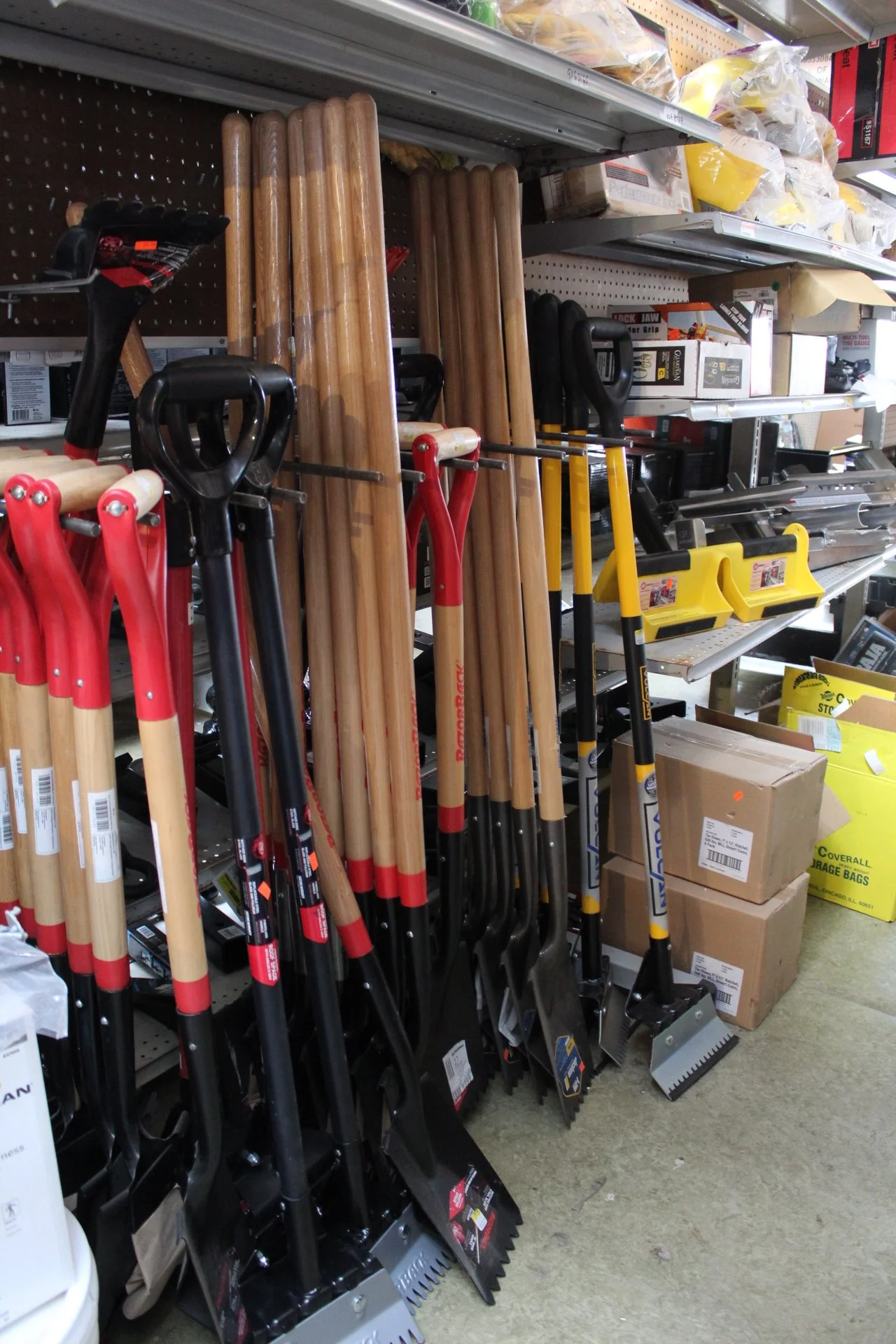 A store shelf displaying various shovels with wooden handles and black or red blades, along with other hardware tools and supplies.