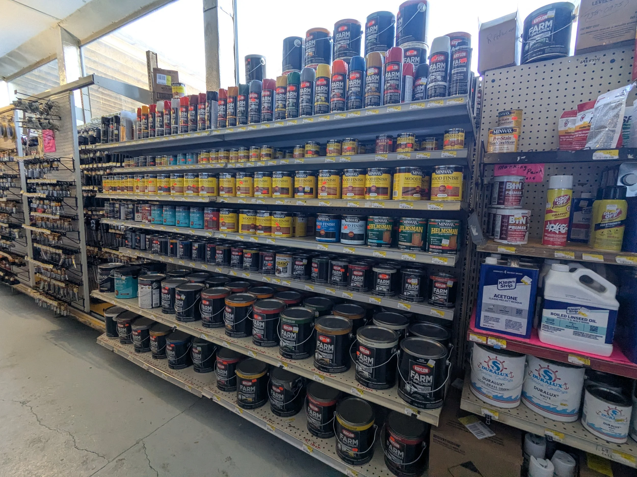 Aisle of hardware store shelves stocked with cans of paint and wood finish, with a side display of primers and cleaning supplies.