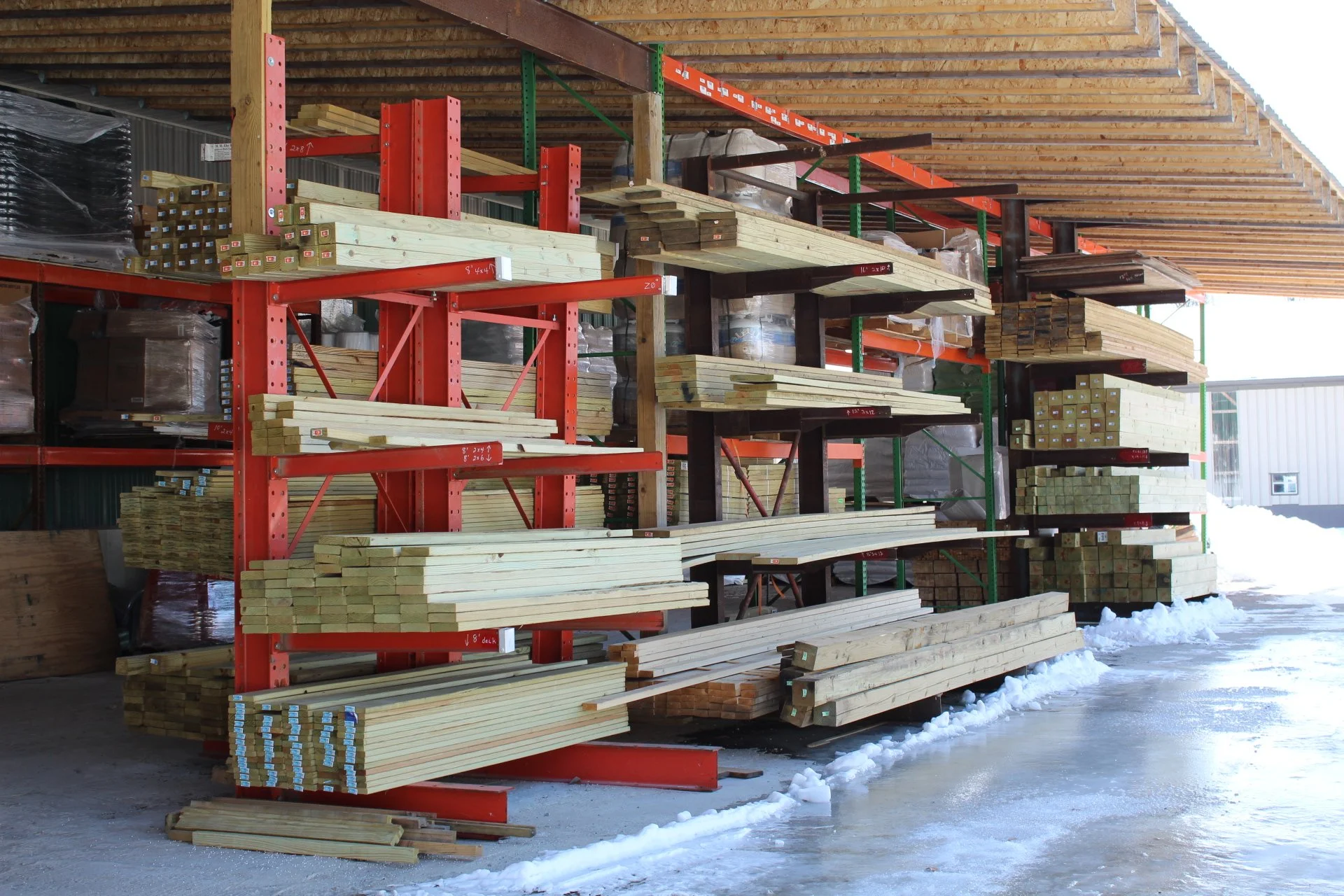 S pring wood stored on outdoor shelves and racks under a wooden roof in a hardware store, with snow on the ground.