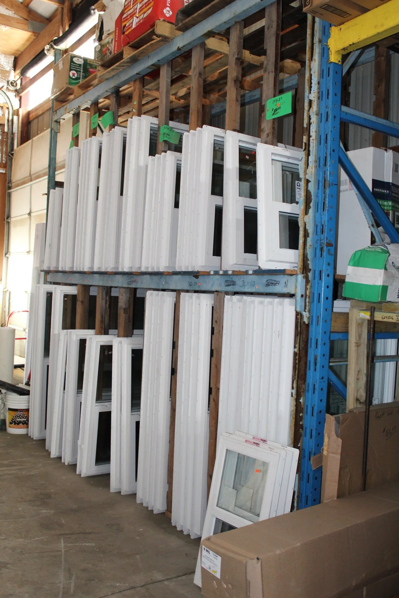 Multiple white window frames stored upright in a warehouse on a metal and wooden shelving unit, with boxes and supplies around.