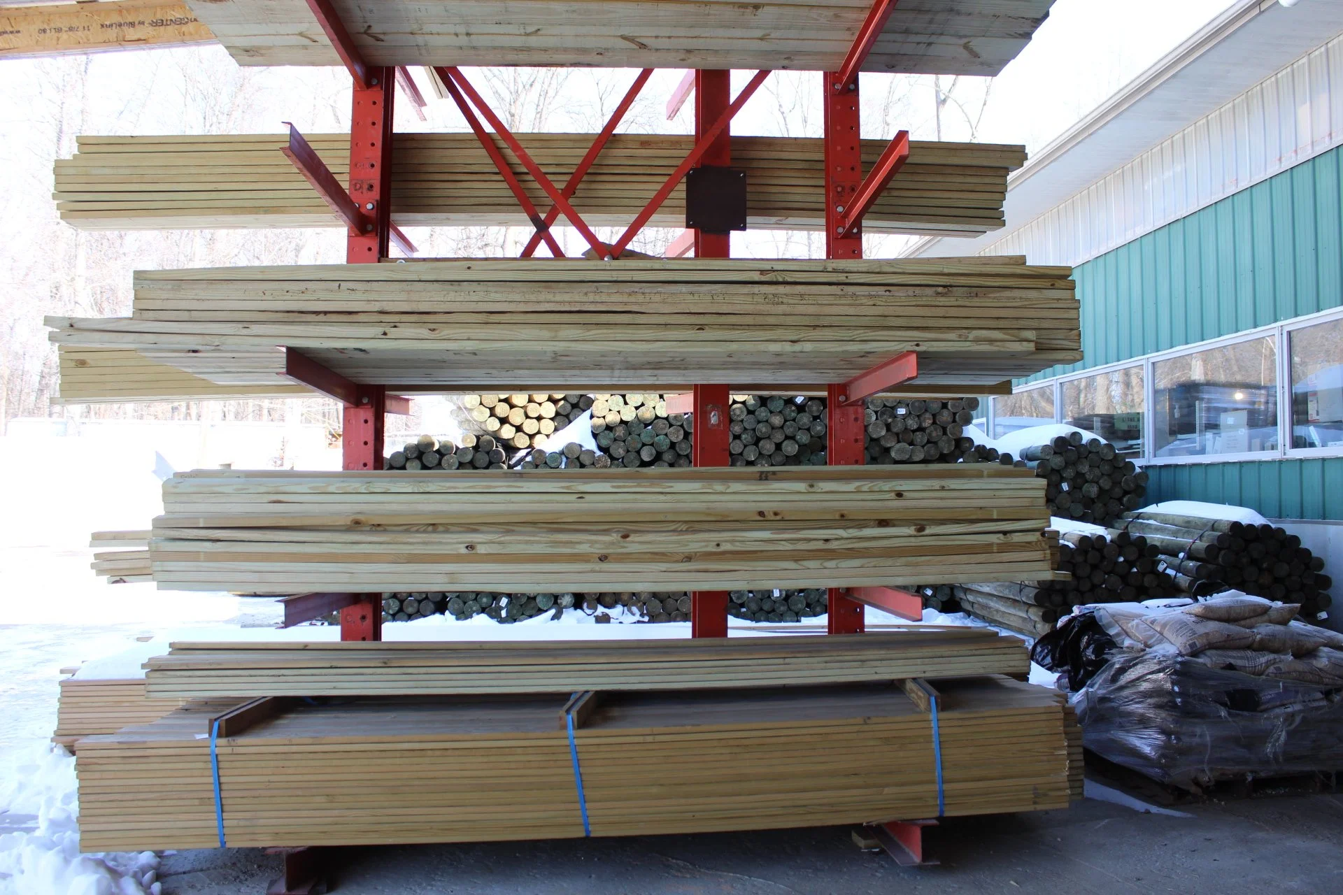 Wood planks stacked on a red metal outdoor storage shelf, with a snow-covered ground and a green building in the background.