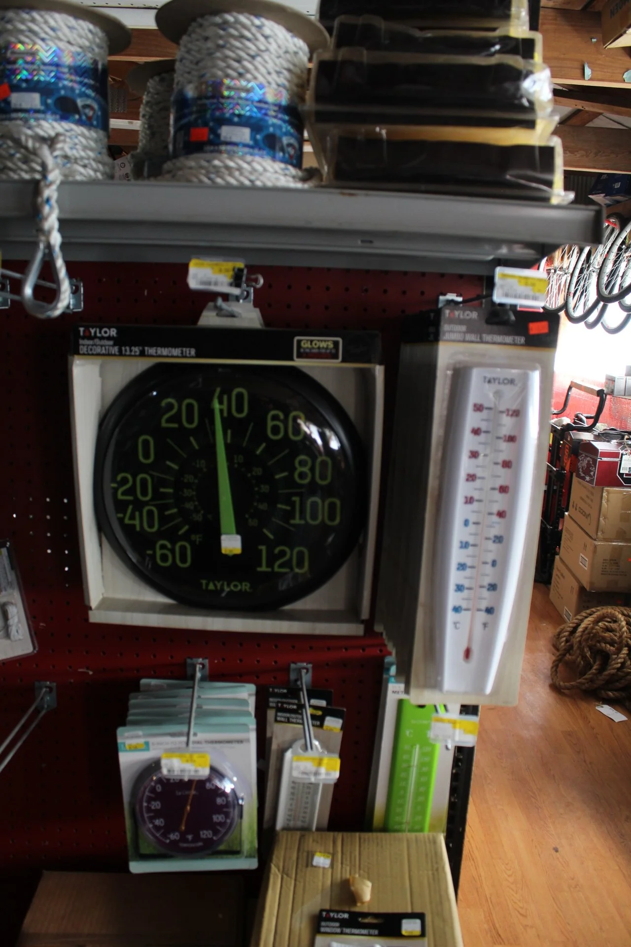 A store display of various thermometers, including a large black indoor/outdoor thermometer showing the temperature around 20°F, a white stick thermometer, and a small round thermometer, all on a red pegboard in a hardware store.