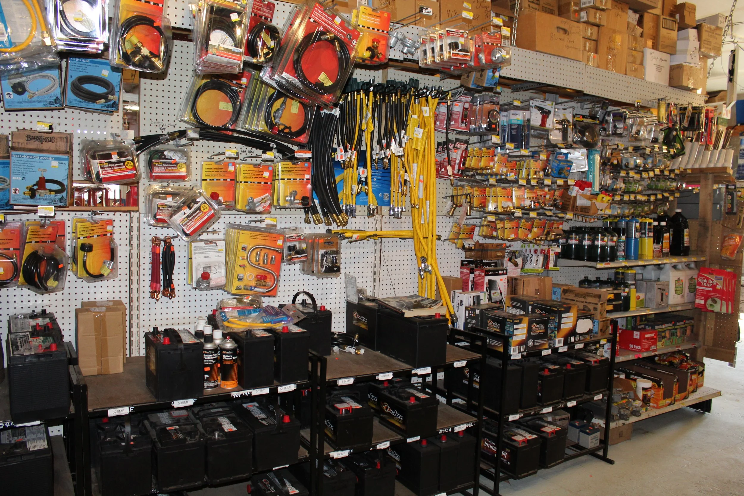 A display of various hardware tools and supplies on wall shelves and on black metal tables in a store. Items include hoses, cables, batteries, and other hardware accessories.