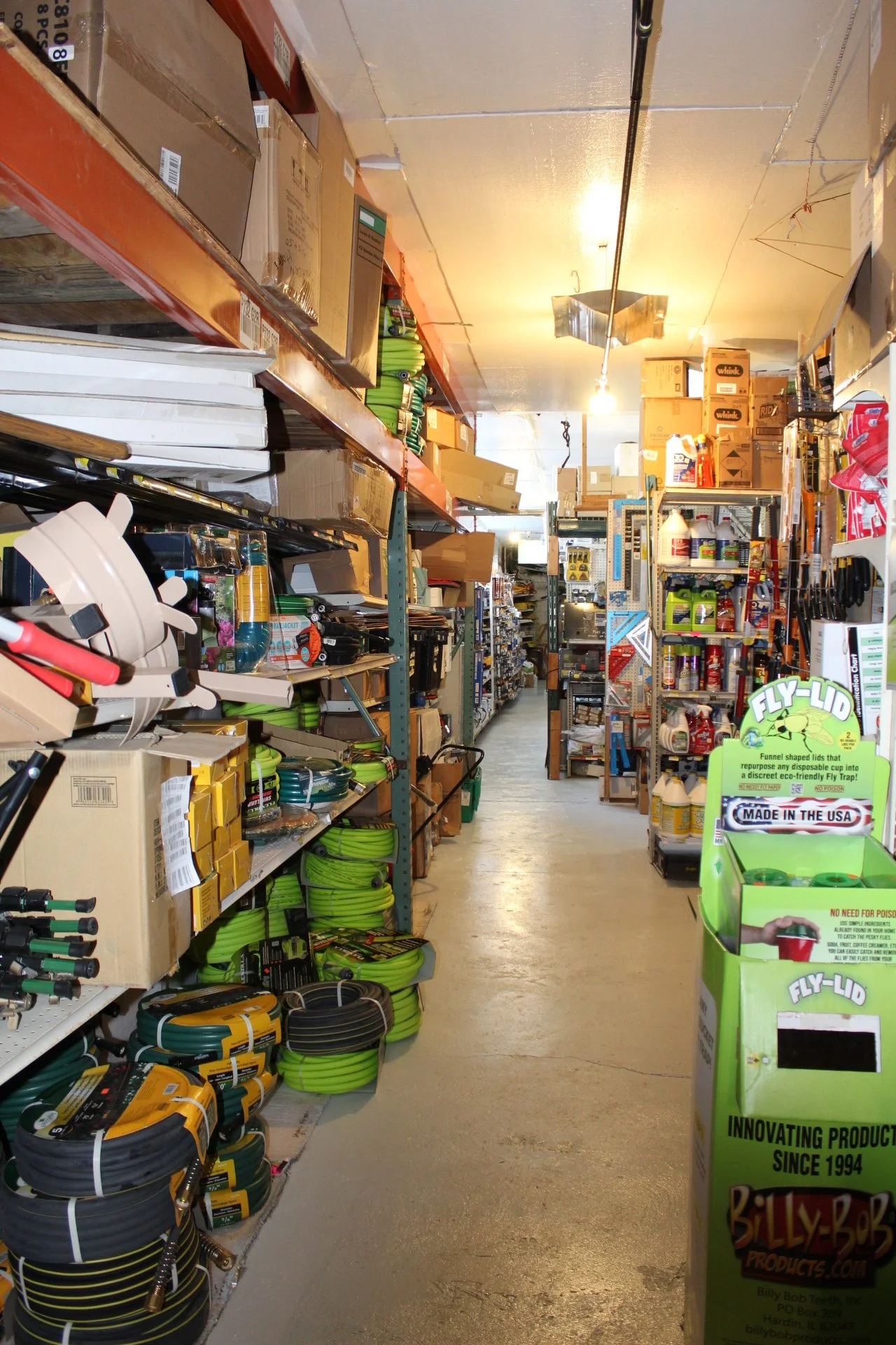 A store aisle with shelves stocked with various gardening and outdoor supplies, including hoses, tools, and chemicals, with a green Billy-Bob fly-lid repurposing product display at the front.