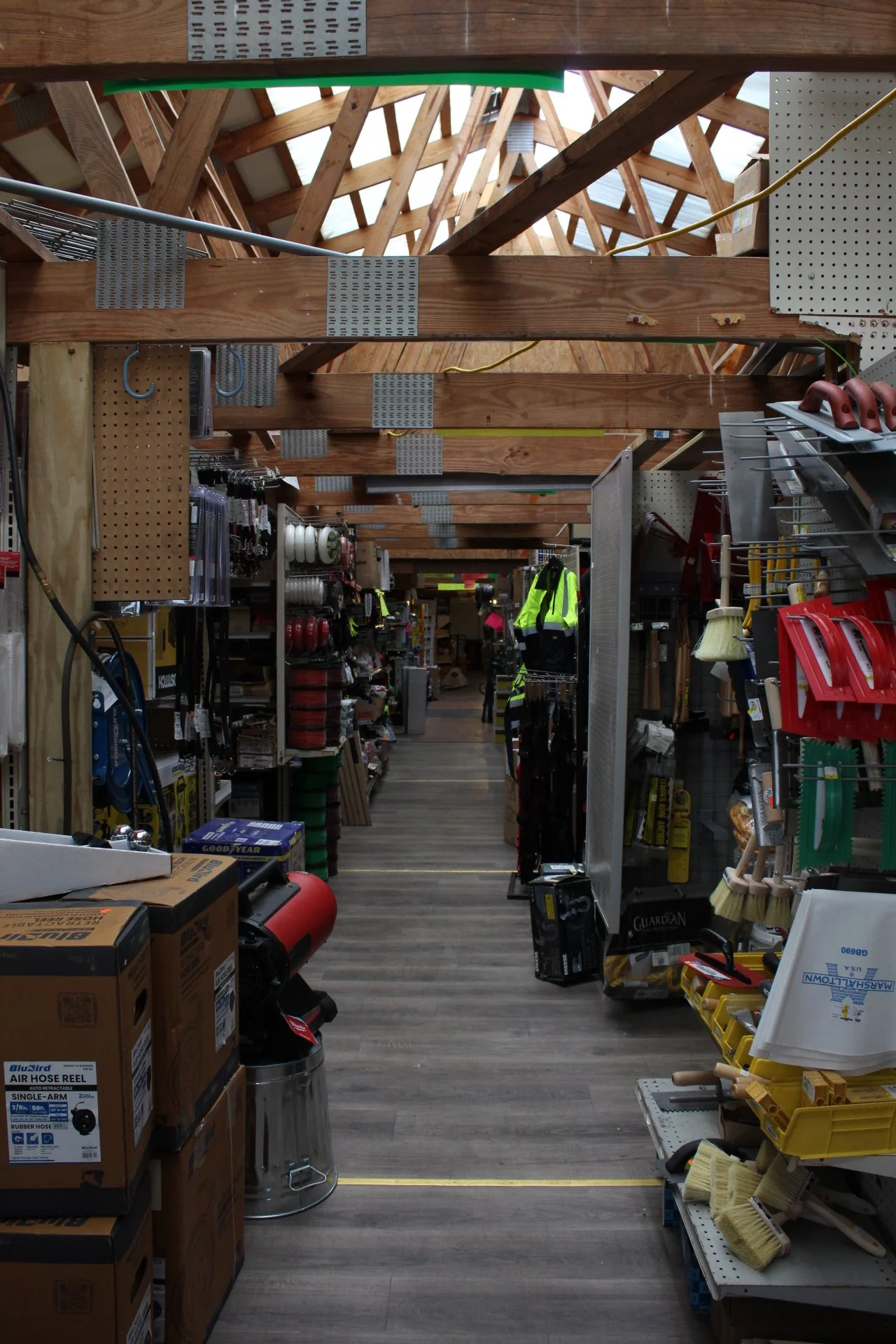 View inside a hardware store aisle with shelves stocked with tools, safety gear, and supplies, under an unfinished wooden roof structure.