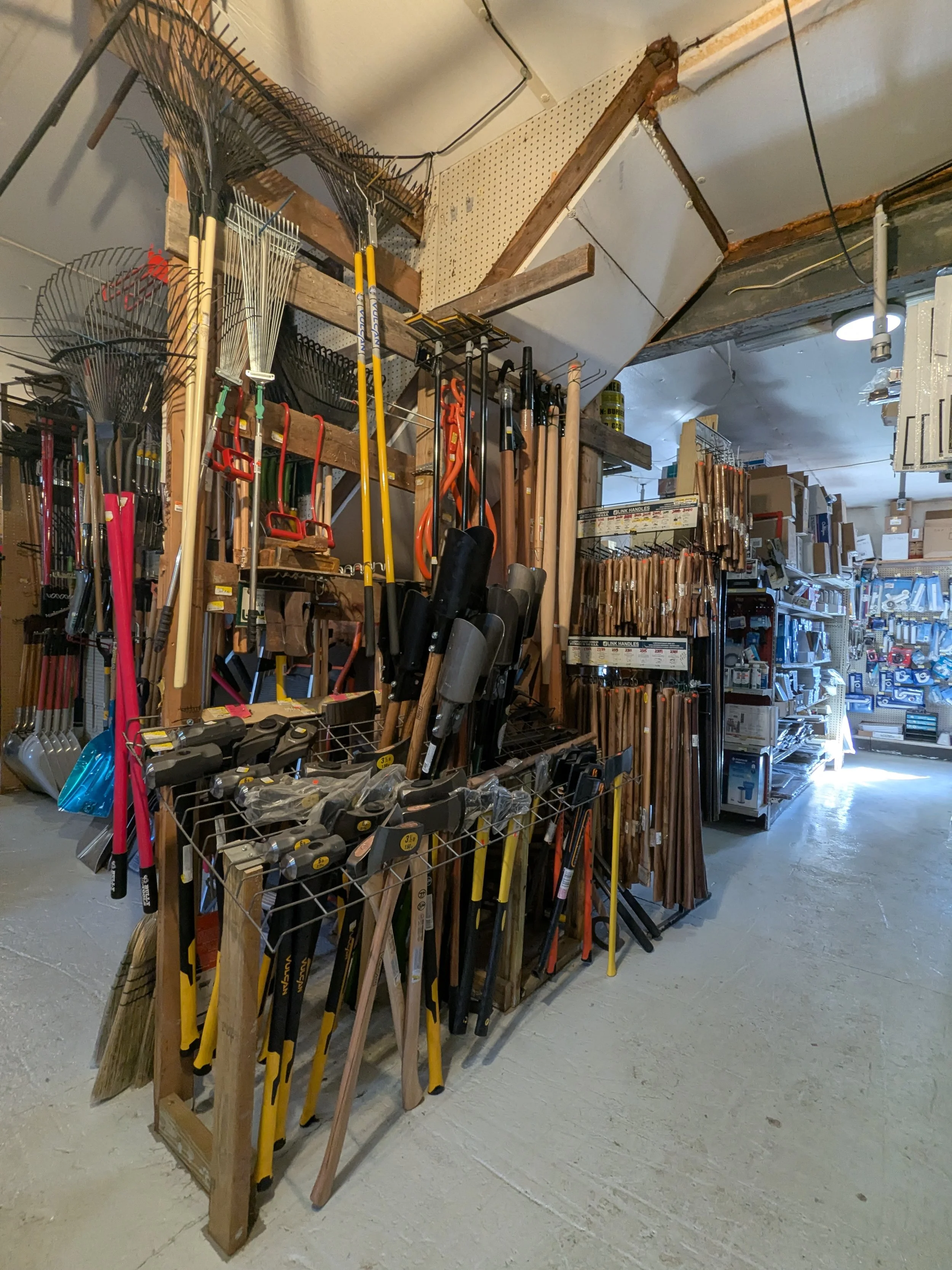 A hardware store aisle with gardening tools and supplies including rakes, shovels, hoes, and other yard tools arranged on wooden shelves and metal racks.
