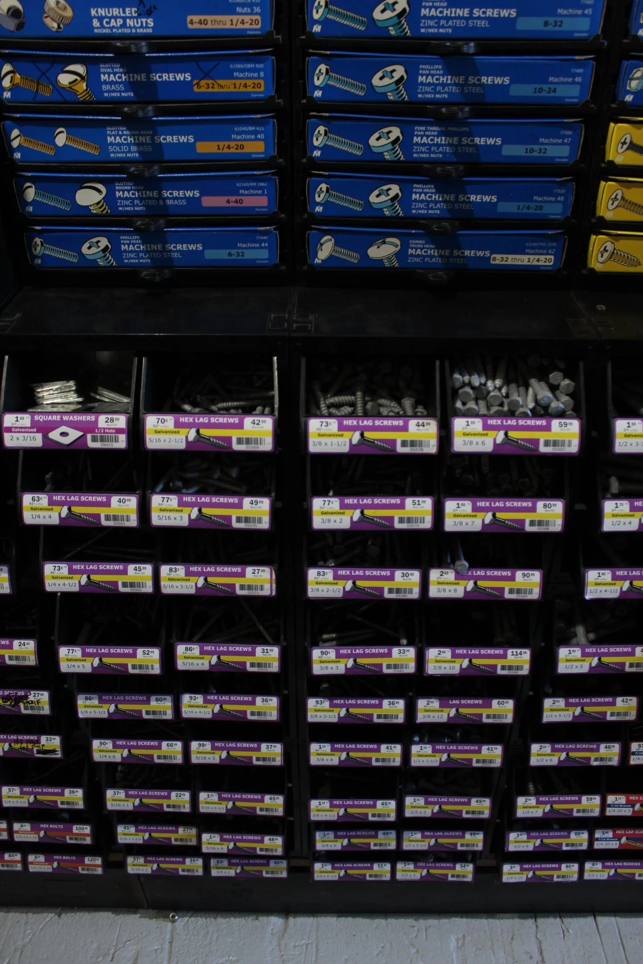 Shelf with various sizes of screws and fasteners in packaging, organized in rows. Top row has blue boxes of machine screws, middle and bottom rows contain small boxes with different types of hex lag screws and other fasteners, with colorful labels di