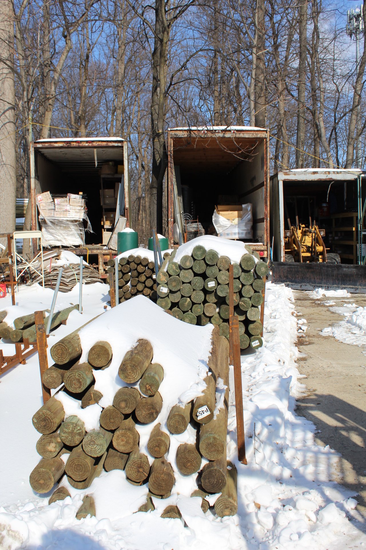 Stacks of cut logs covered in snow in front of open truck storage units filled with various supplies and equipment, with a snow-covered ground and leafless trees in the background.