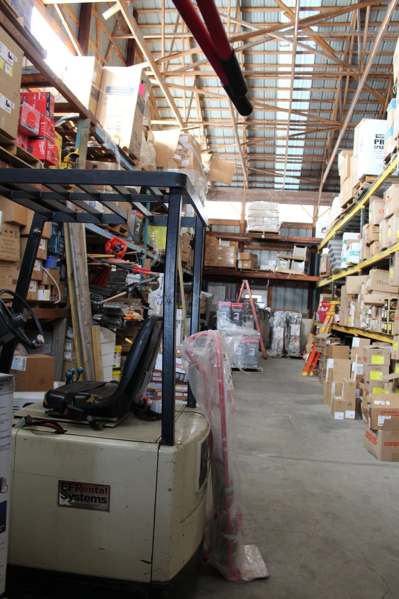 View of a storage warehouse with high metal roof and wooden beams, filled with shelves of boxes, pallets, and equipment, including a forklift in the foreground.