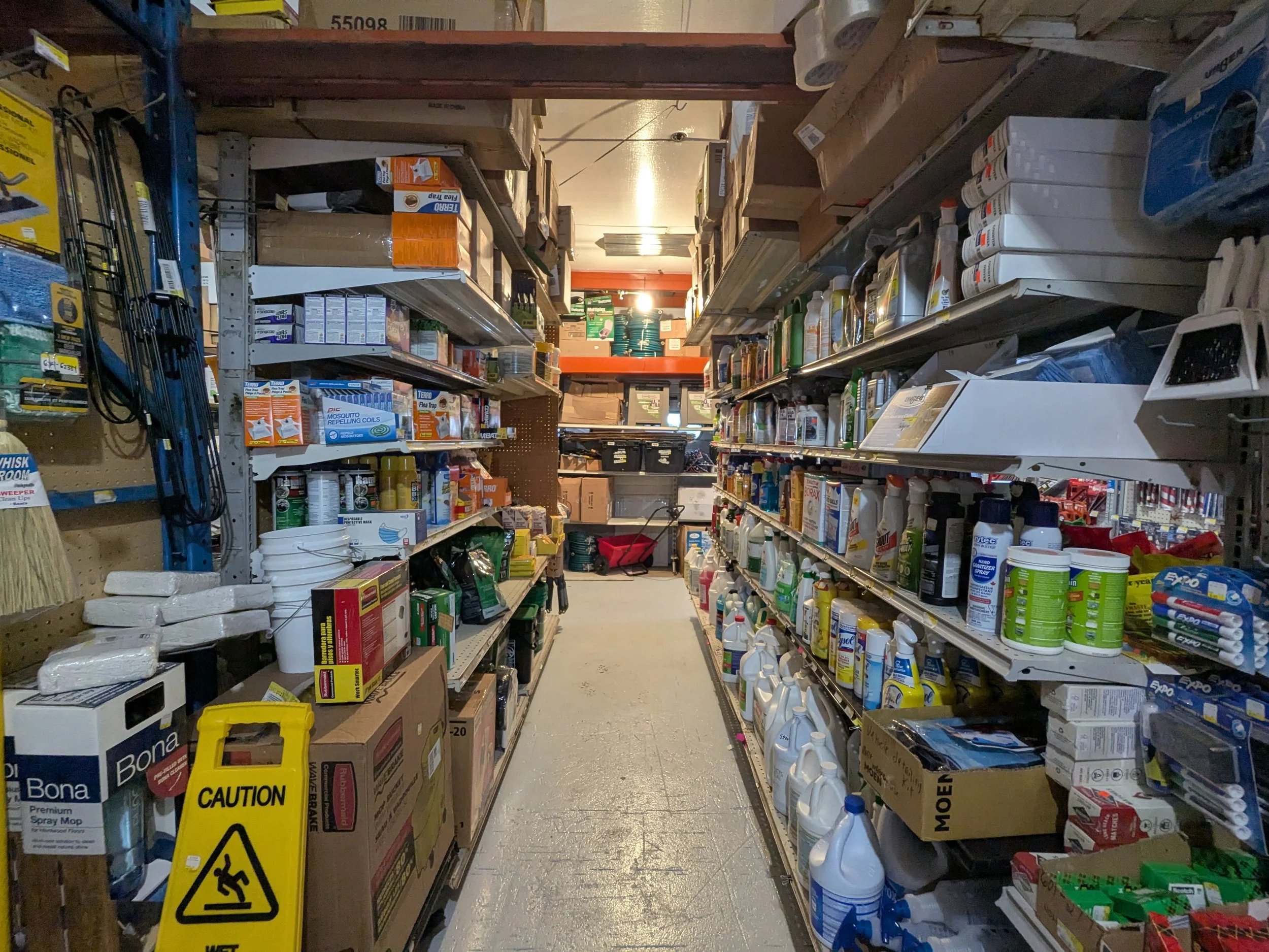 A store aisle with shelves stocked with various household supplies, including cleaning products, sprays, bottles, and hardware tools, with a wet floor caution sign in the foreground.