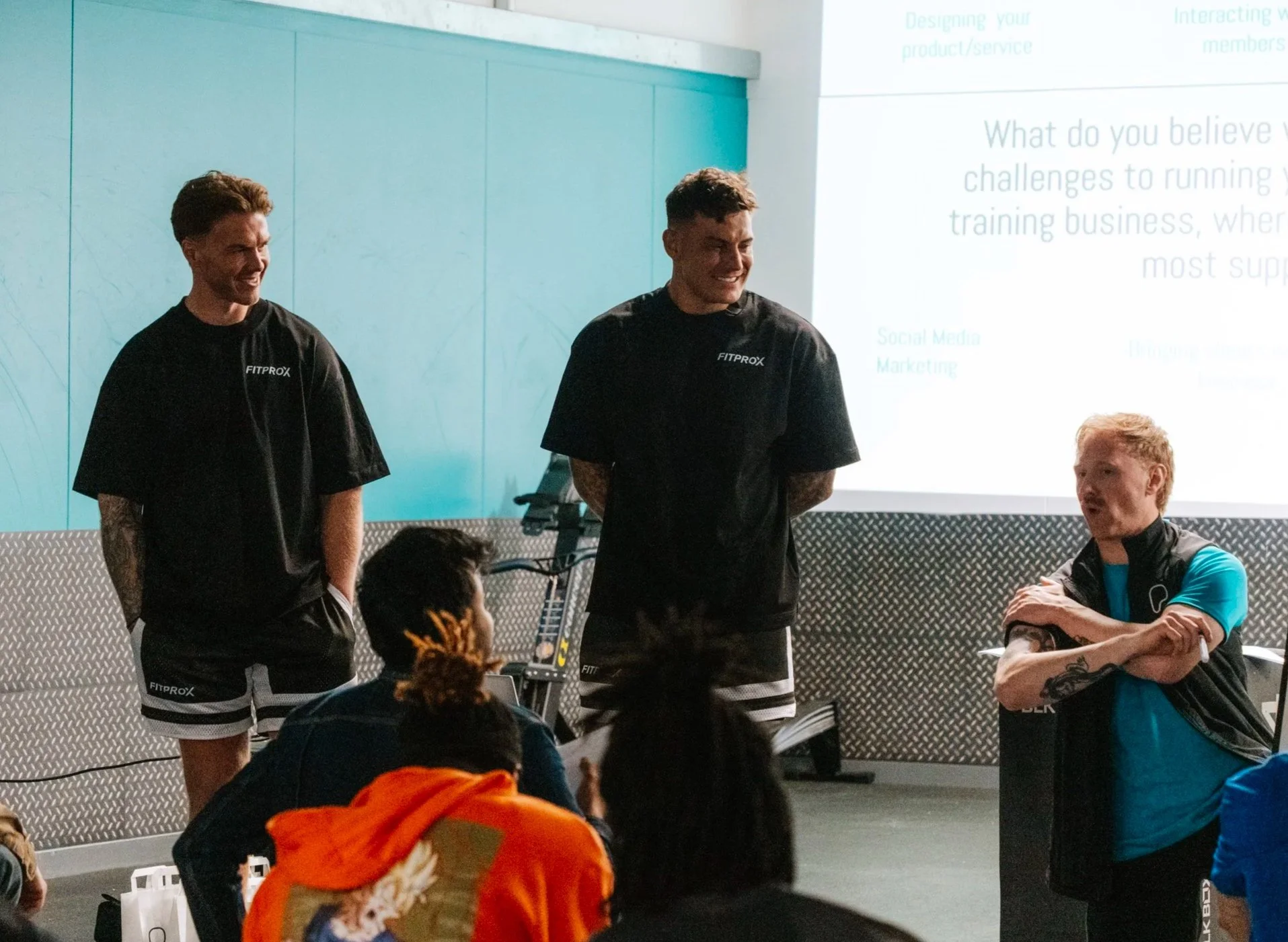 Three men standing and talking in front of a group of people in a fitness or training room. One man is sitting and speaking to the group. There is a projection on the wall.