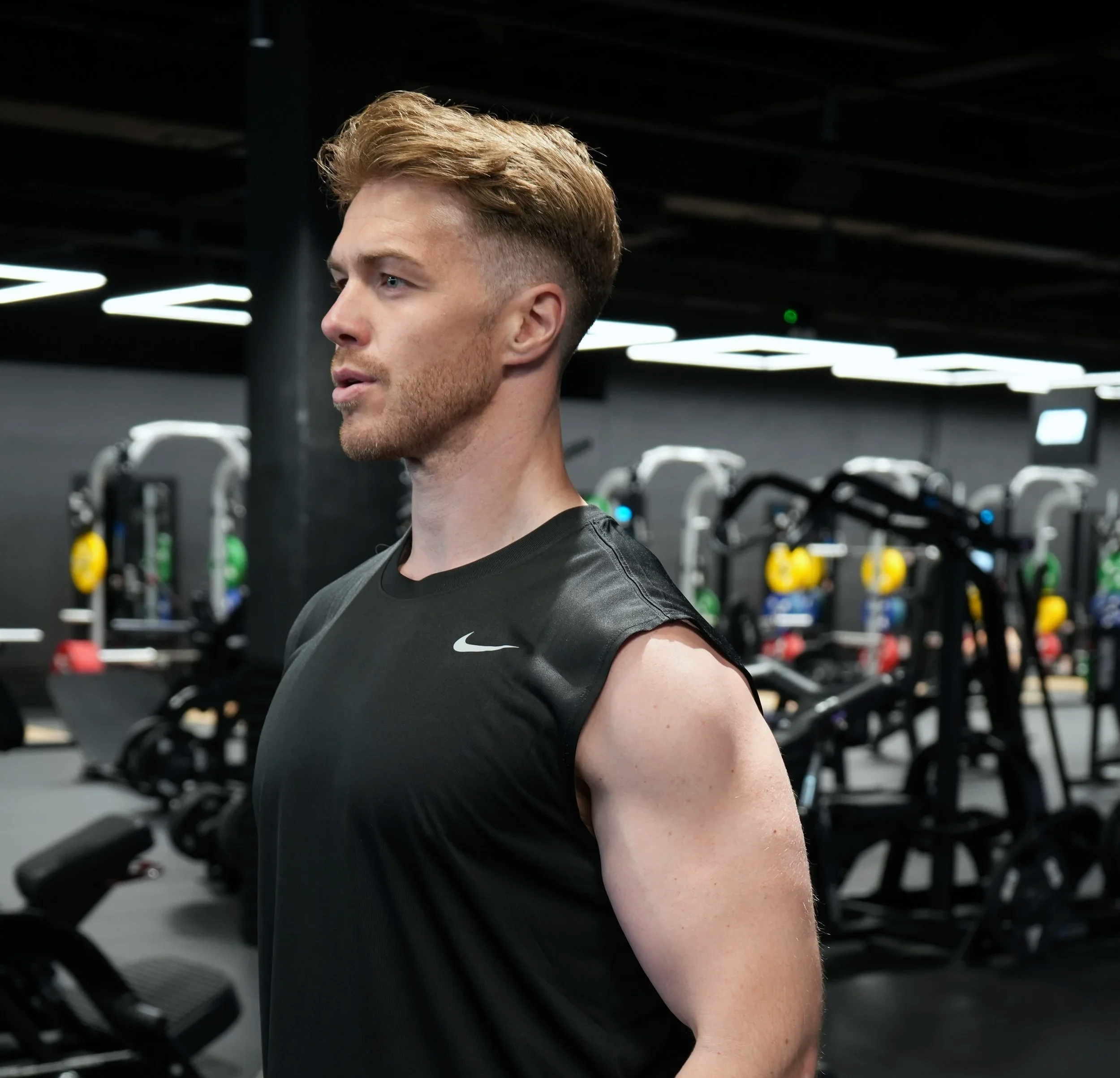 Man in a black sleeveless Nike shirt at a gym with exercise equipment in the background.