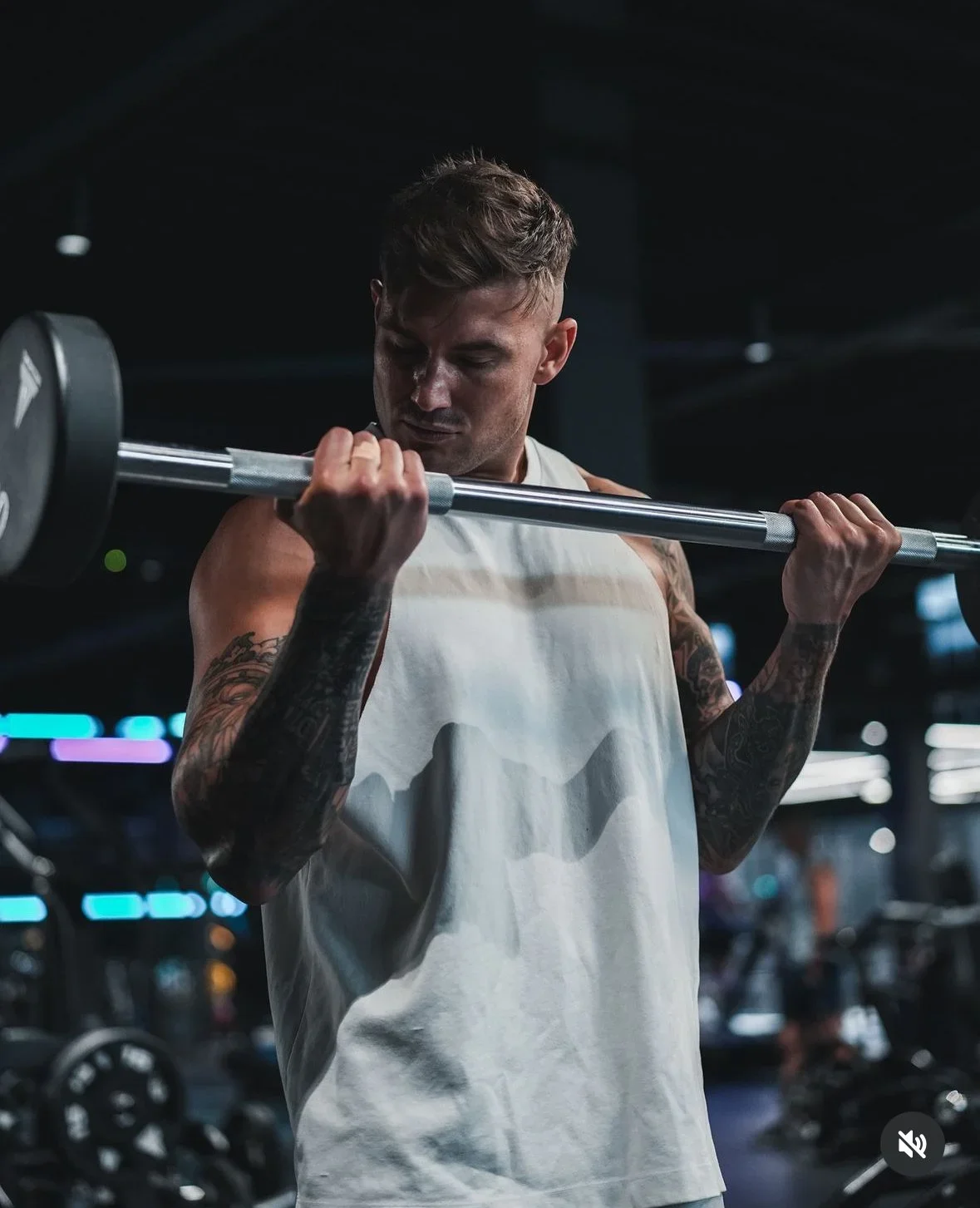 Man lifting a barbell in a gym with machines and lighting in the background.