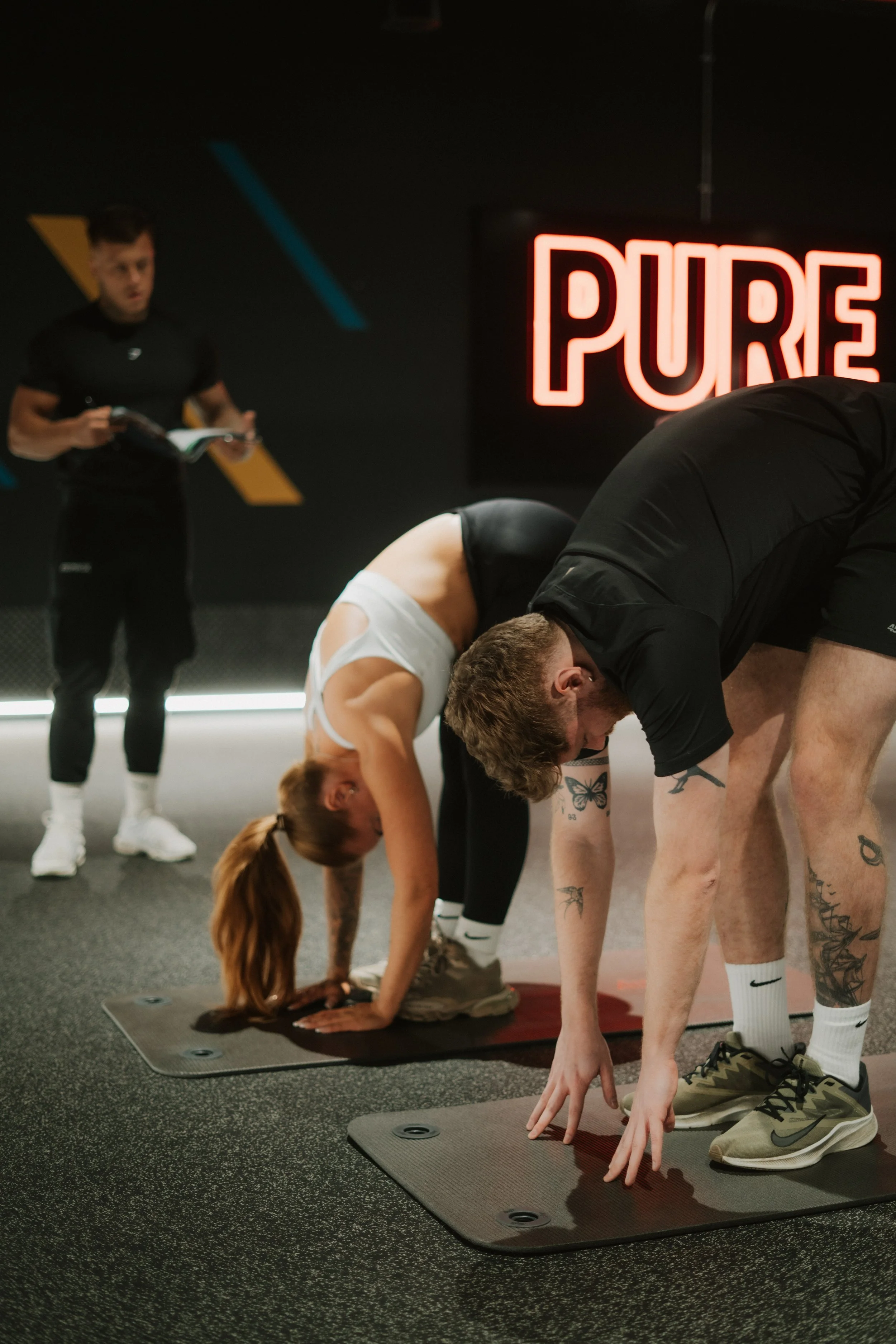 Group of people doing yoga stretches on mats in a gym with a neon 'PURE' sign on the wall.