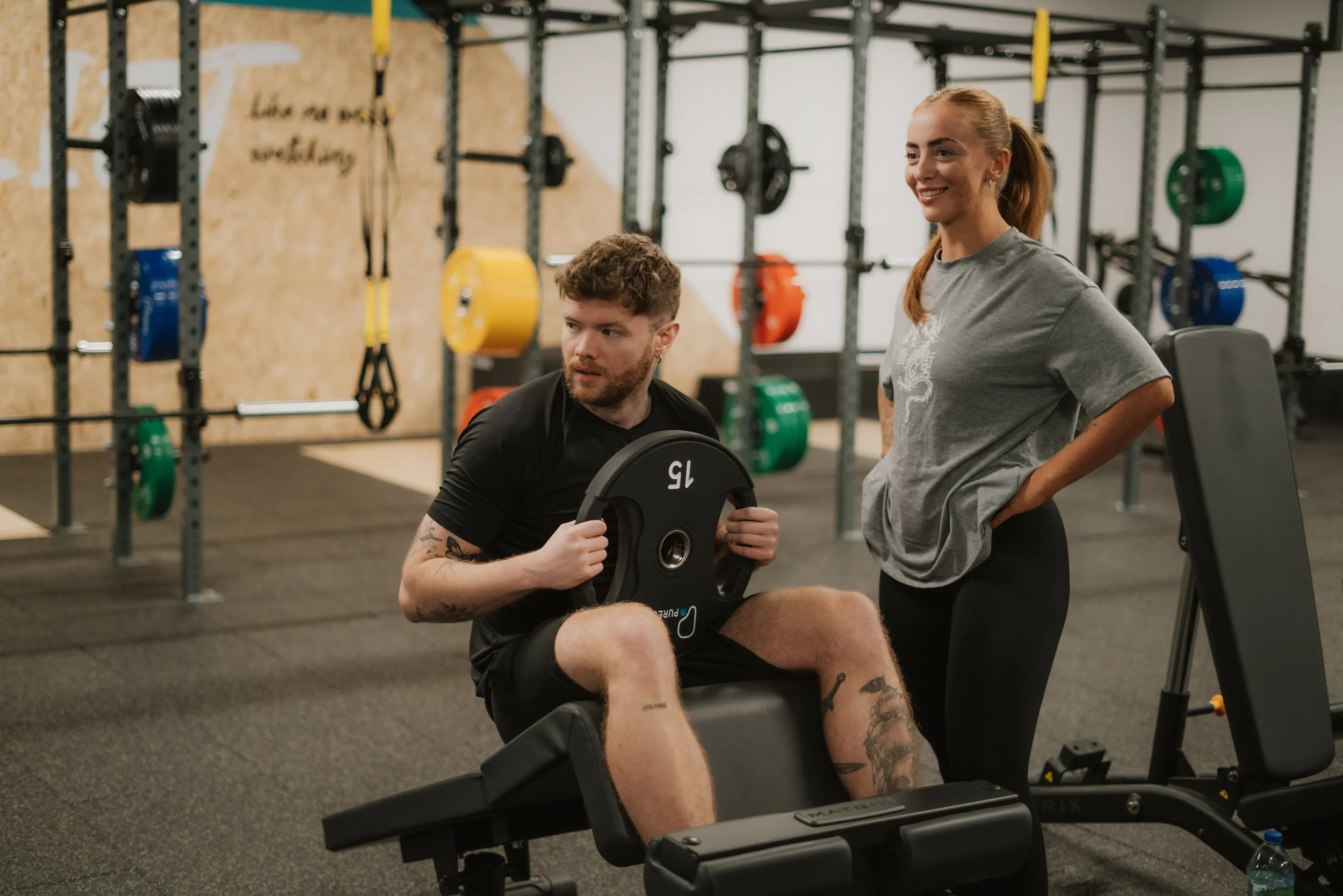 A man sitting on a workout bench holding a weight plate, with a woman standing next to him in a gym filled with colorful weights and equipment.
