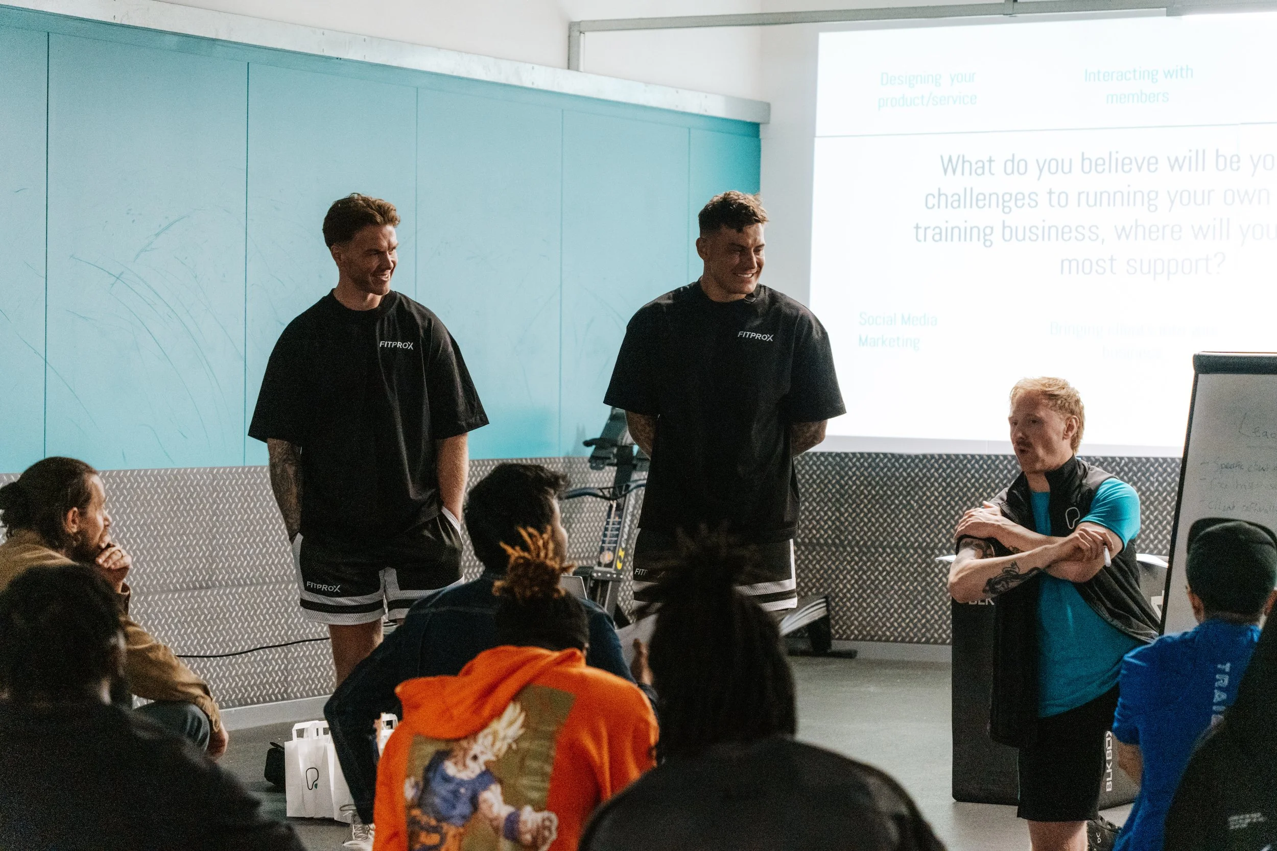 Group of people attending a fitness workshop or seminar. Two men in black shirts stand at the front, leading the discussion, while others sit and listen. Presentation slide projected on the wall behind.