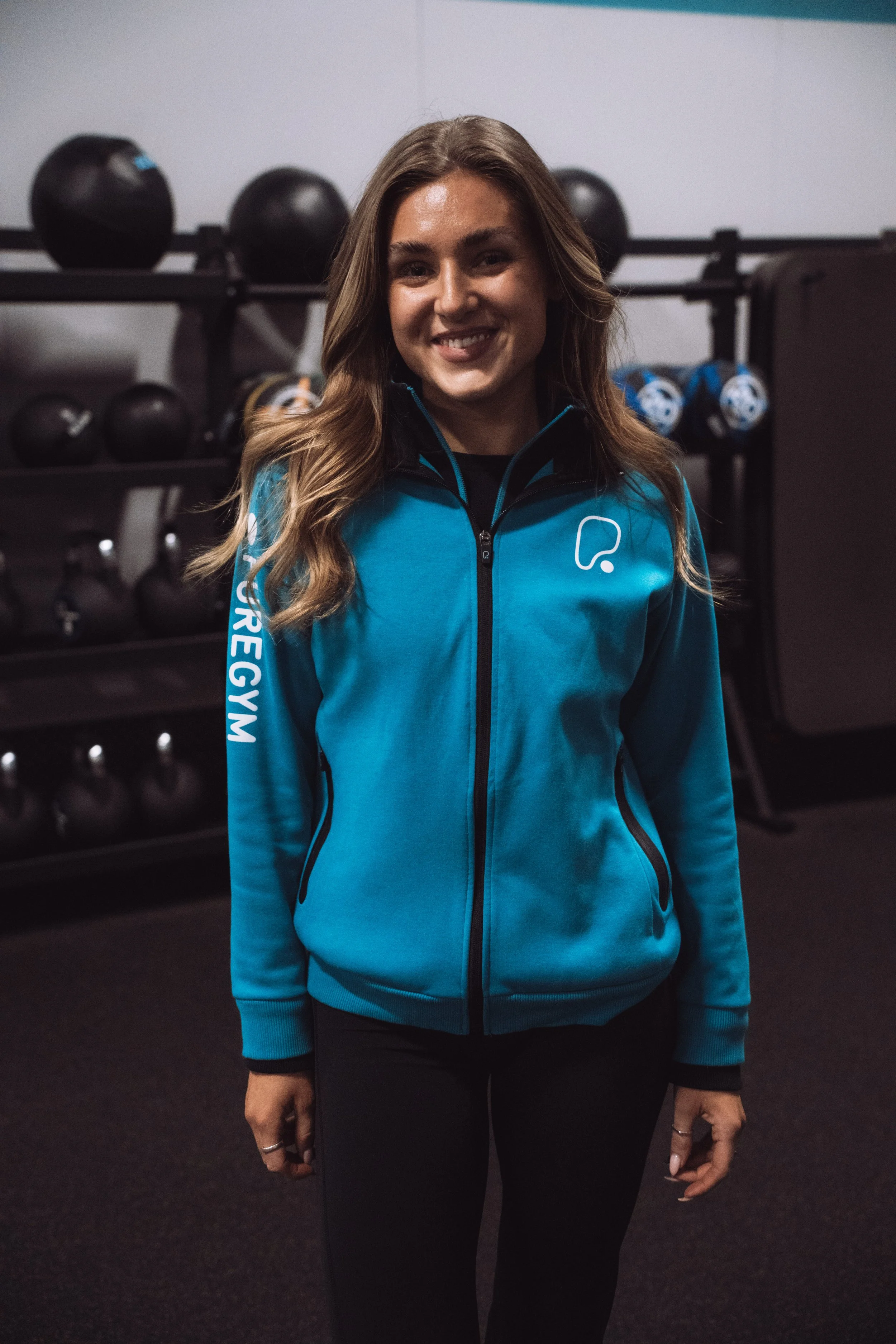 A woman in a blue jacket standing in a gym with kettlebells in the background.