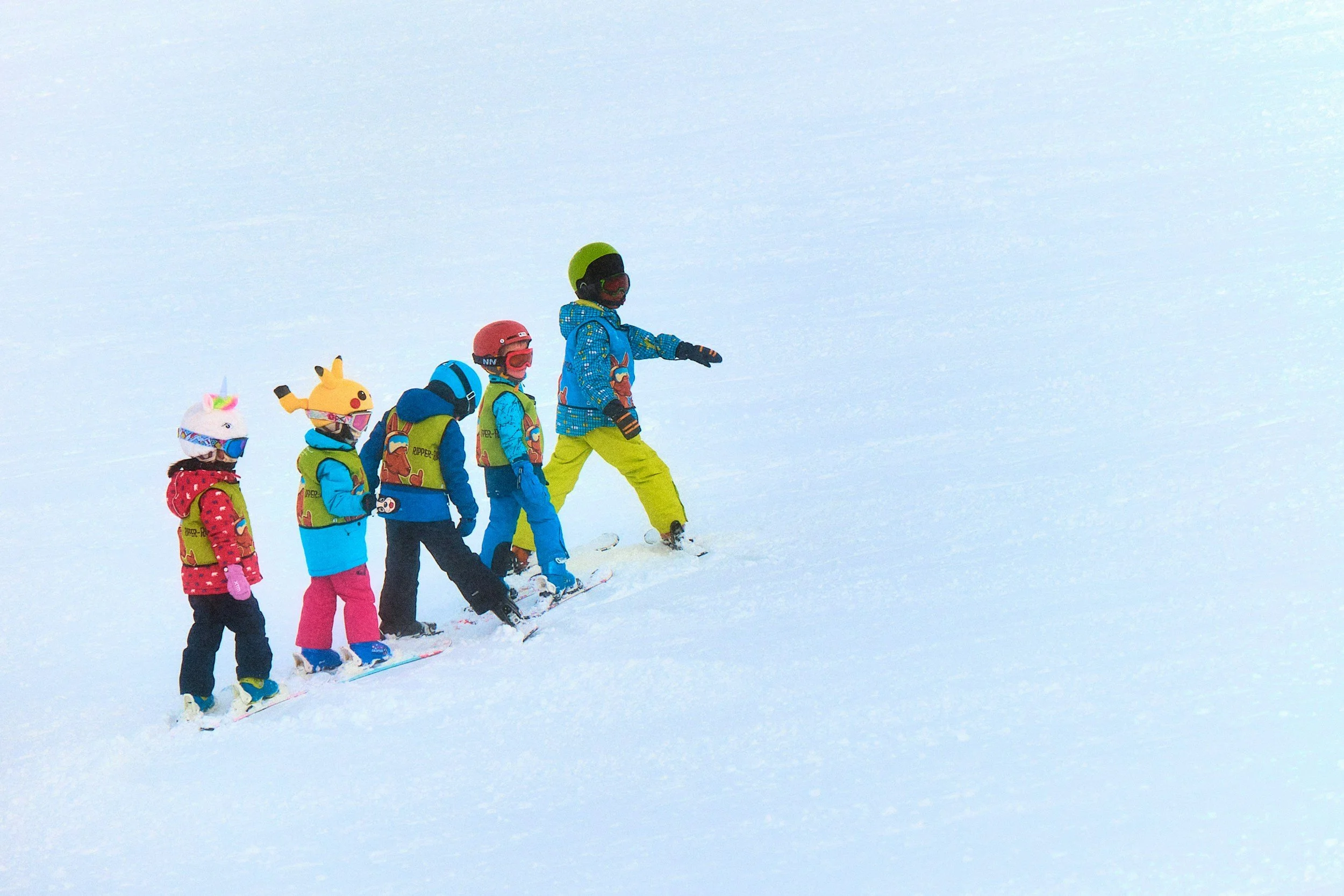 Group of children learning to ski on snow-covered slope, with an instructor guiding them.